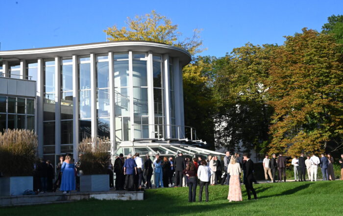 Absolventinnen und Absolventen in festlicher Kleidung versammeln sich vor einem modernen Glasgebäude, umgeben von grünem Rasen und Bäumen unter strahlend blauem Himmel – ein unvergesslicher Abschied. at Hochschule Coburg