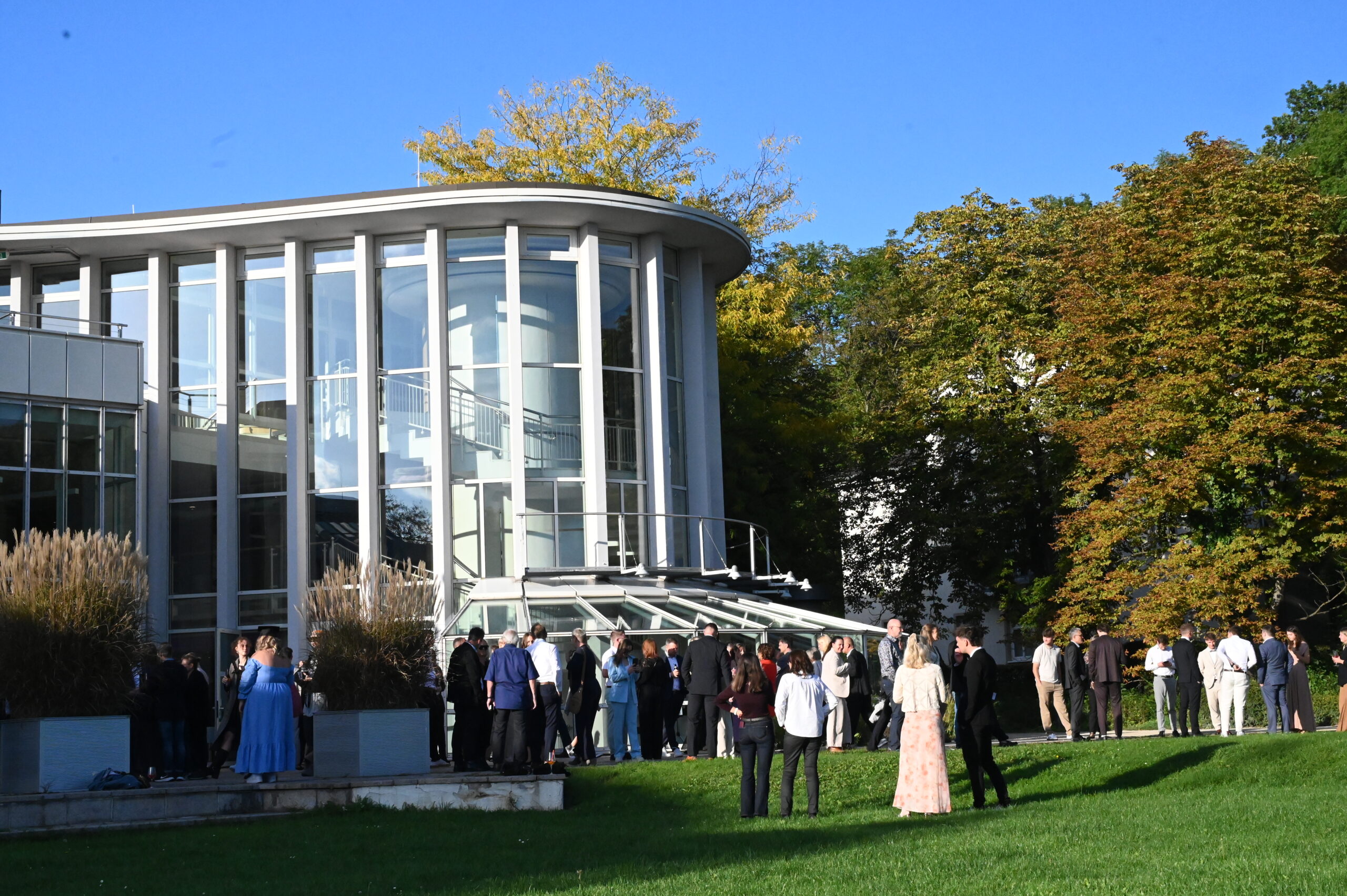 Absolventinnen und Absolventen in festlicher Kleidung versammeln sich vor einem modernen Glasgebäude, umgeben von grünem Rasen und Bäumen unter strahlend blauem Himmel – ein unvergesslicher Abschied. at Hochschule Coburg
