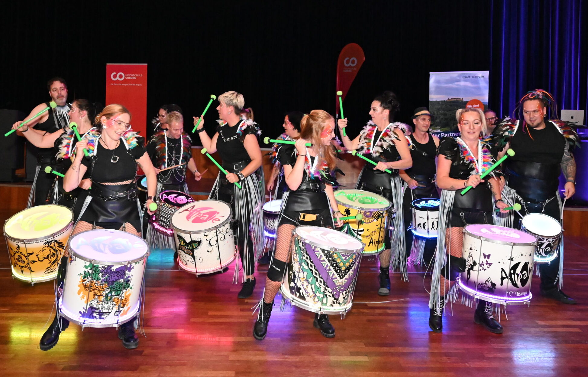 Eine Gruppe von Darstellern, die den Abschied der Absolventinnen und Absolventen feiert, spielt auf einer Bühne mit Holzboden und dunklen Vorhängen auf großen, bunten Trommeln mit grünen Trommelstöcken. at Hochschule Coburg