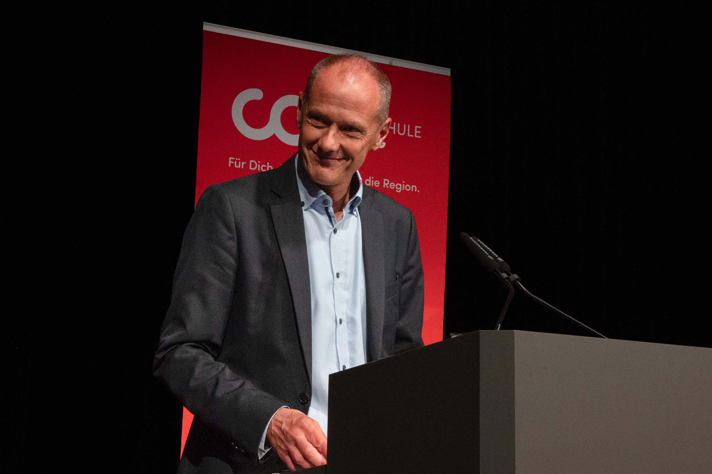 Ein Mann im Anzug steht lächelnd am Rednerpult, im Hintergrund sind ein Mikrofon und ein rotes Schild mit einem deutschen Text über Absolventinnen und Absolventen zu sehen. at Hochschule Coburg