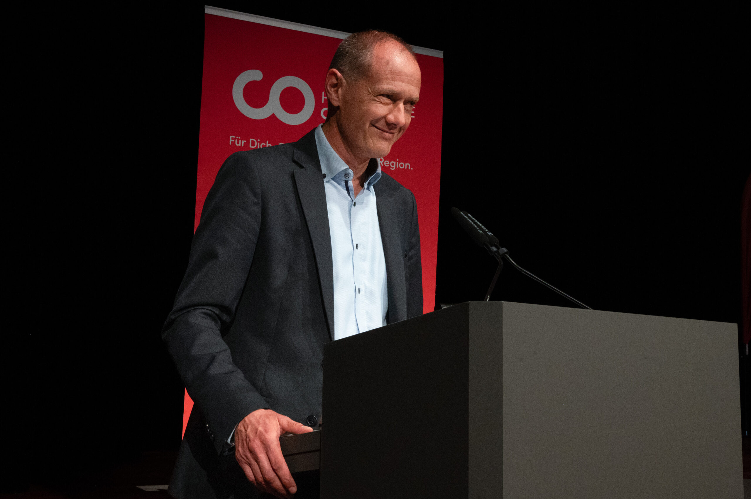 Ein Mann im Anzug steht am Rednerpult und spricht in ein Mikrofon. Hinter ihm ist ein rotes Schild mit weißer Schrift und einem Logo zu sehen, während er sich bei der Abschiedsfeier an die Absolventinnen und Absolventen wendet. at Hochschule Coburg