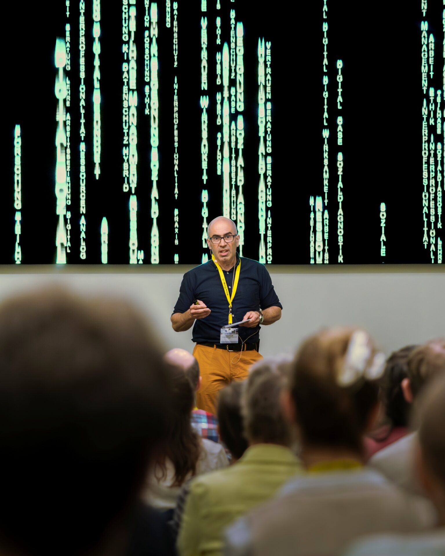 Ein Mann hält vor einem Publikum einen Vortrag und steht dabei vor einer Leinwand, auf der grüner Code zu sehen ist, ähnlich wie im Film „Matrix“. at Hochschule Coburg