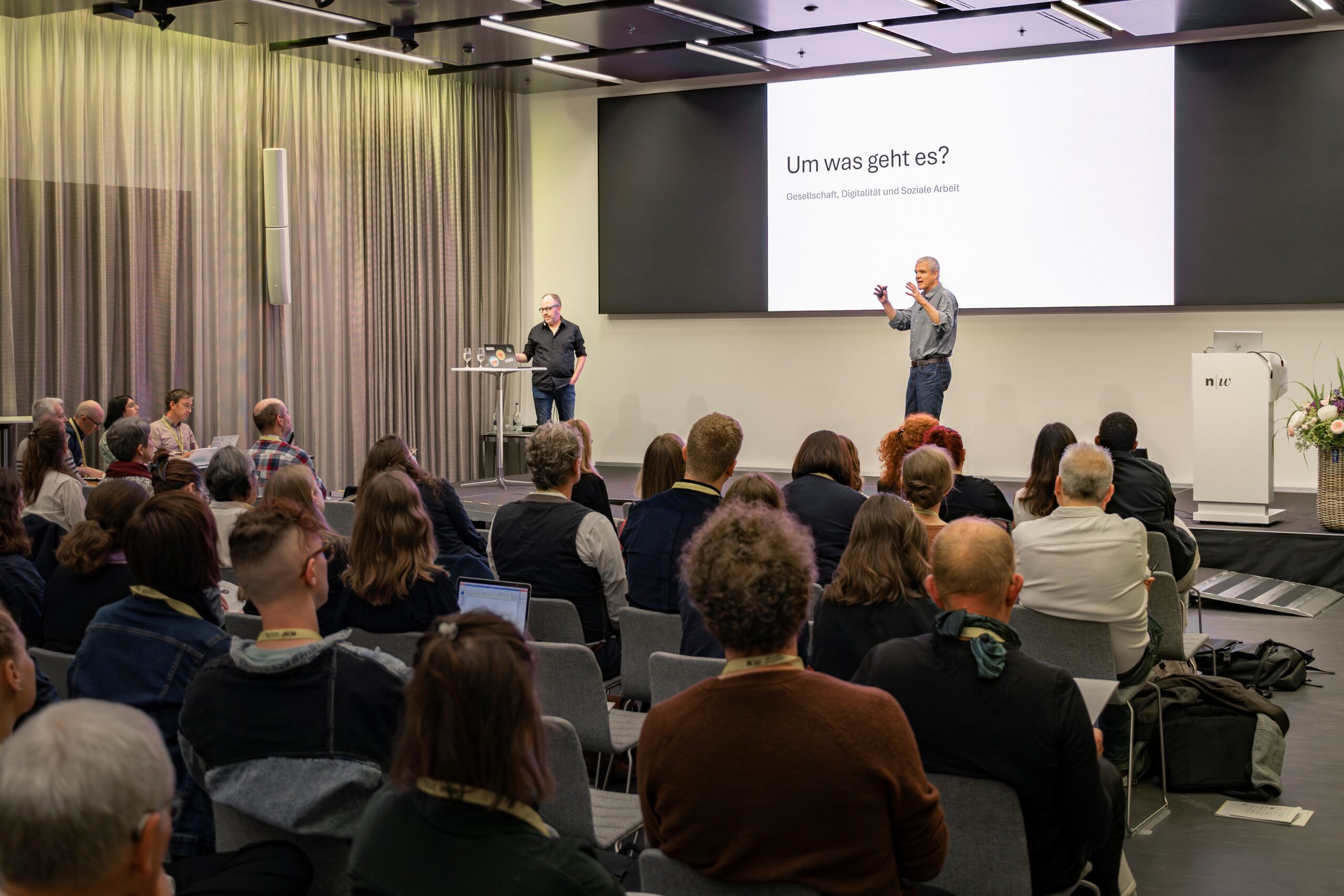 Ein Redner spricht in einem modernen Konferenzraum zu den Zuhörern, während eine andere Person neben einem Laptop steht. Auf einem Bildschirm hinter ihnen ist ein deutscher Text zu sehen: „Um was geht es?“, zusammen mit einem Untertitel. at Hochschule Coburg