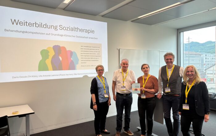 Fünf Personen stehen lächelnd in einem hellen Klassenzimmer, hinter ihnen ist eine Präsentationsfolie mit dem Titel „Weiterbildung Sozialtherapie“ zu sehen. Durch das Fenster auf der rechten Seite sind ein Berg und Gebäude zu erkennen. at Hochschule Coburg