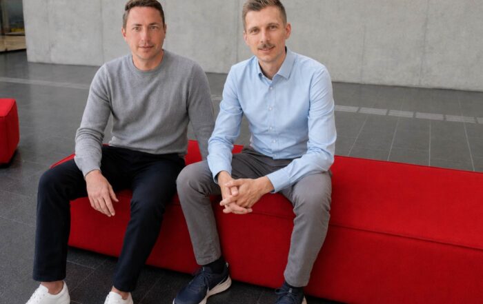 Zwei Männer sitzen drinnen auf einer leuchtend roten Bank. Der eine trägt einen grauen Pullover und eine schwarze Hose, der andere ein hellblaues Hemd und eine graue Hose. Im Hintergrund ist eine schlichte, hellgraue Wand zu sehen – ein passend zurückhaltender Rahmen für Tim Brasch, dessen jüngste ESG-Forschung internationale Auszeichnungen erhalten hat. at Hochschule Coburg