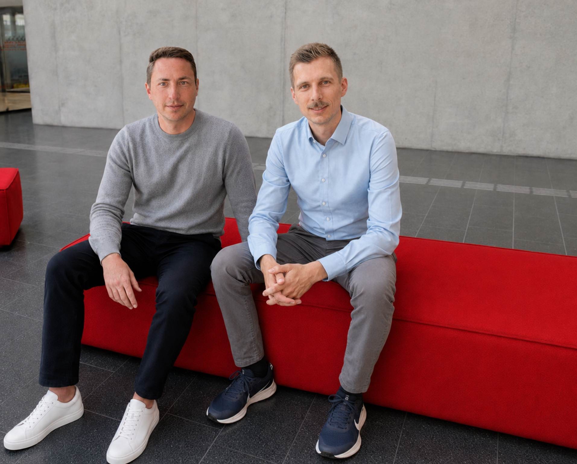Zwei Männer sitzen drinnen auf einer leuchtend roten Bank. Der eine trägt einen grauen Pullover und eine schwarze Hose, der andere ein hellblaues Hemd und eine graue Hose. Im Hintergrund ist eine schlichte, hellgraue Wand zu sehen – ein passend zurückhaltender Rahmen für Tim Brasch, dessen jüngste ESG-Forschung internationale Auszeichnungen erhalten hat. at Hochschule Coburg