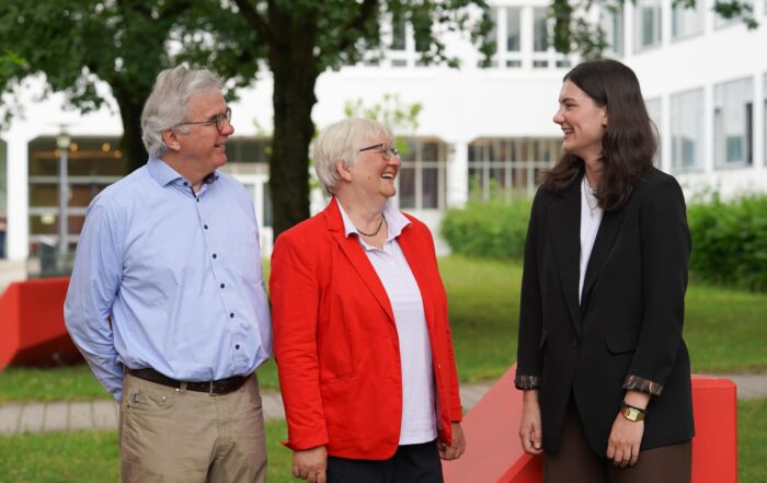 Drei Menschen stehen draußen, lächeln und unterhalten sich. Zwei ältere Erwachsene – einer in einem roten Blazer und einer in einem blauen Hemd – stehen einer jüngeren Frau in einem schwarzen Blazer gegenüber. Ihr freundlicher Austausch spiegelt wider, wie engagierte Gespräche Chancen eröffnen und dabei helfen, gemeinsam die Zukunft zu gestalten. Im Hintergrund sind Grünflächen und ein weißes Gebäude zu sehen. at Hochschule Coburg