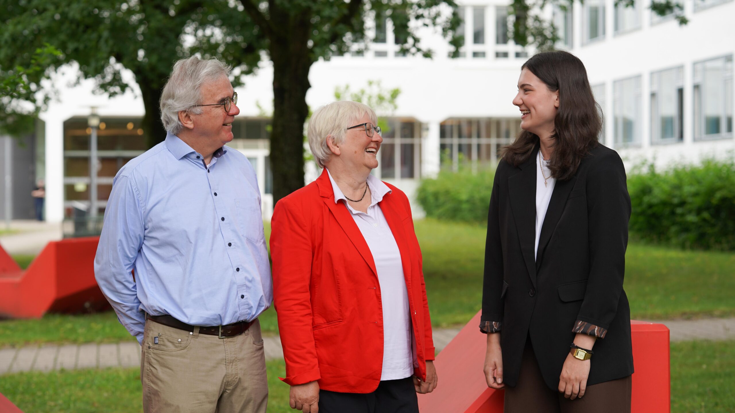 Drei Menschen stehen draußen, lächeln und unterhalten sich. Zwei ältere Erwachsene – einer in einem roten Blazer und einer in einem blauen Hemd – stehen einer jüngeren Frau in einem schwarzen Blazer gegenüber. Ihr freundlicher Austausch spiegelt wider, wie engagierte Gespräche Chancen eröffnen und dabei helfen, gemeinsam die Zukunft zu gestalten. Im Hintergrund sind Grünflächen und ein weißes Gebäude zu sehen. at Hochschule Coburg