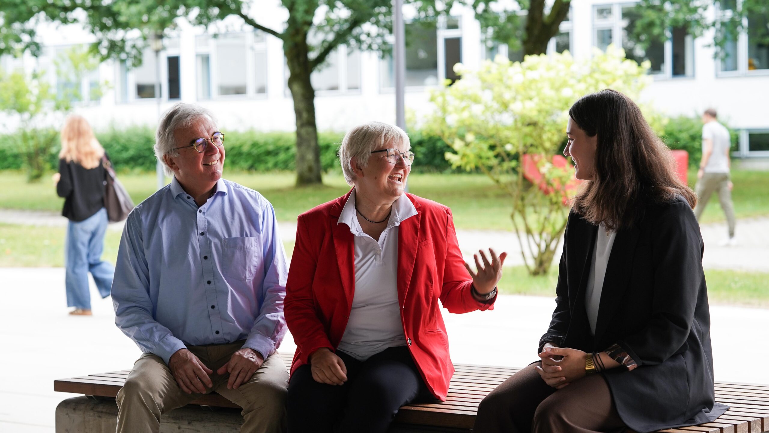 Drei Menschen sitzen auf einer Bank im Freien, unterhalten sich und lächeln. Ihre lebhafte Diskussion spiegelt ihr gemeinsames Engagement für „Zukunft gestalten“ und „Engagement fördern“ wider. Im Hintergrund sind Bäume, Sträucher und ein verschwommenes Gebäude mit Fenstern zu sehen. at Hochschule Coburg