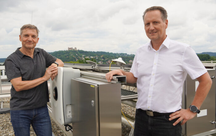 Zwei Männer stehen auf einem Dach neben großen wissenschaftlichen Geräten, die innovative Ansätze für die Energieversorgung der Zukunft symbolisieren. Beide blicken in die Kamera, der eine trägt ein graues Hemd, der andere ein weißes. Im Hintergrund ragen Hügel und eine Burg unter einem bewölkten Himmel empor und deuten auf Fortschritt vor dem Hintergrund der Geschichte hin. at Hochschule Coburg