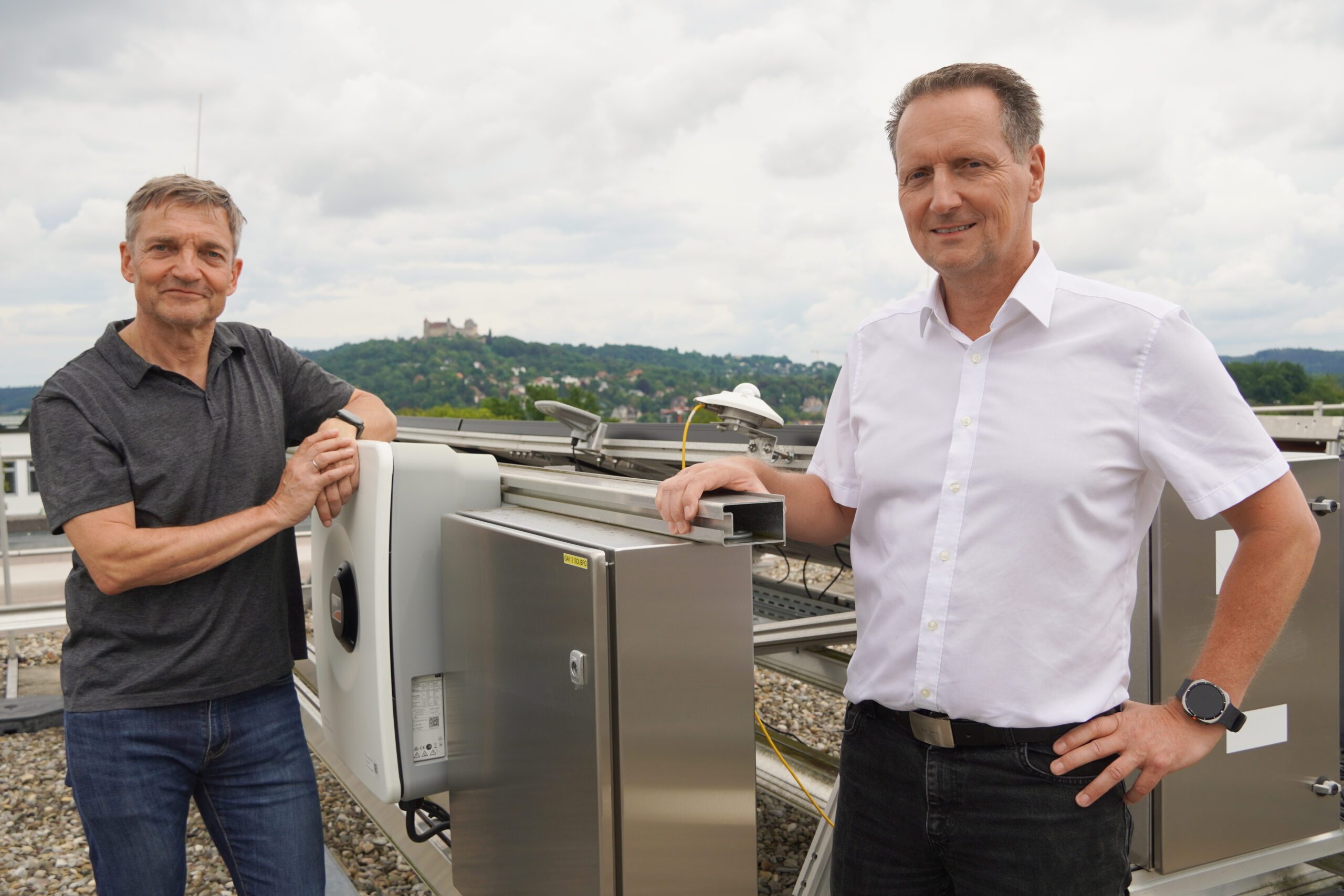 Zwei Männer stehen auf einem Dach neben großen wissenschaftlichen Geräten, die innovative Ansätze für die Energieversorgung der Zukunft symbolisieren. Beide blicken in die Kamera, der eine trägt ein graues Hemd, der andere ein weißes. Im Hintergrund ragen Hügel und eine Burg unter einem bewölkten Himmel empor und deuten auf Fortschritt vor dem Hintergrund der Geschichte hin. at Hochschule Coburg