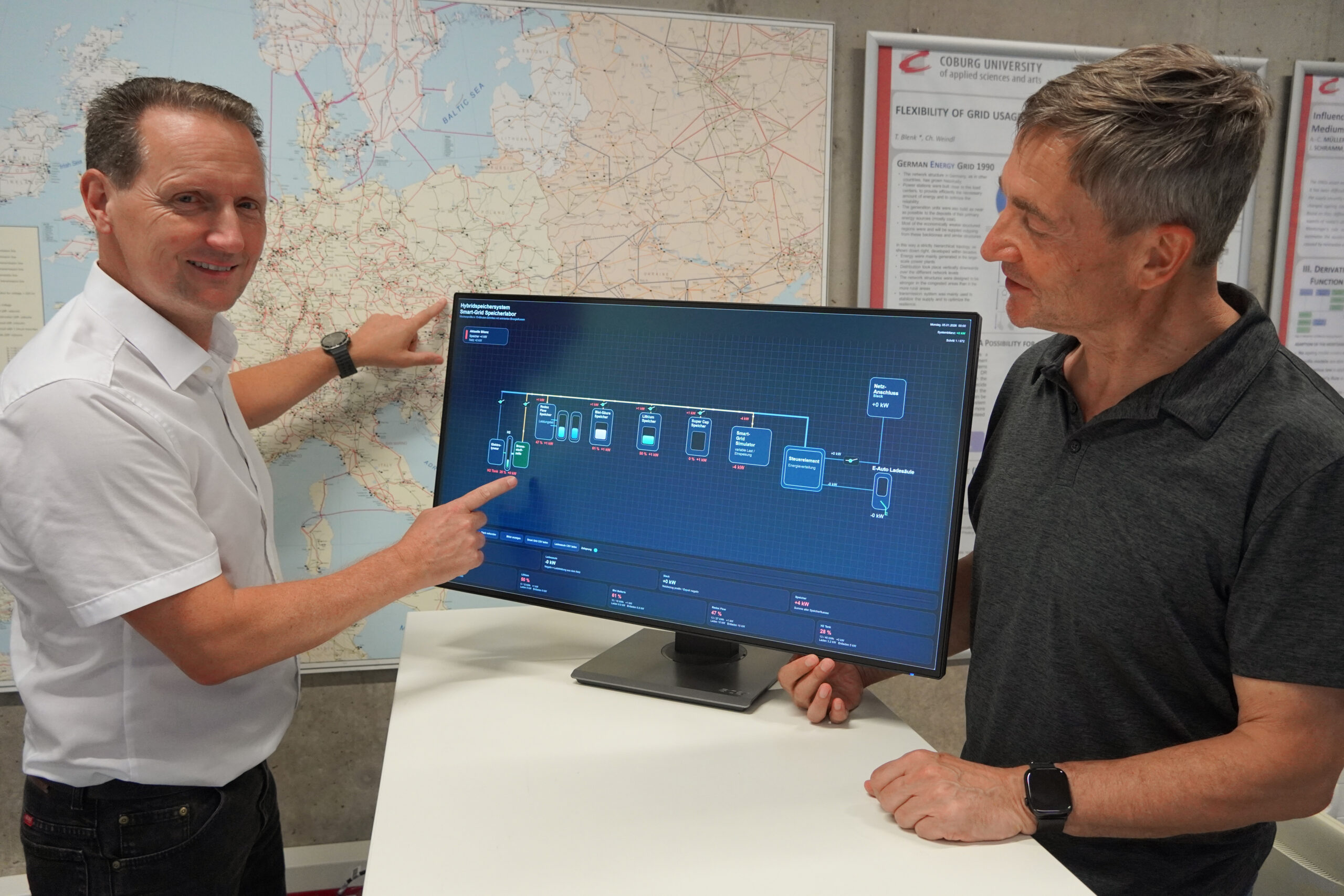 Zwei Männer stehen vor einem Monitor, auf dem ein Flussdiagramm zum Thema Energieversorgung zu sehen ist. Ein Mann zeigt auf den Bildschirm, während der andere nachdenklich zusieht; sie diskutieren über innovative Lösungen für die Zukunft. An der Wand hinter ihnen sind eine große Karte und Plakate zu sehen. at Hochschule Coburg