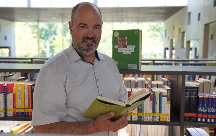 Ein bärtiger Mann steht in einer Bibliothek der Hochschule Coburg, hält ein aufgeschlagenes Buch in der Hand und lächelt. Umgeben von Bücherregalen voller bunter Bücher zu Themen wie Gesundheitswissenschaften und Digital Health genießt er das warme Sonnenlicht, das durch die großen Fenster hereinströmt. at Hochschule Coburg