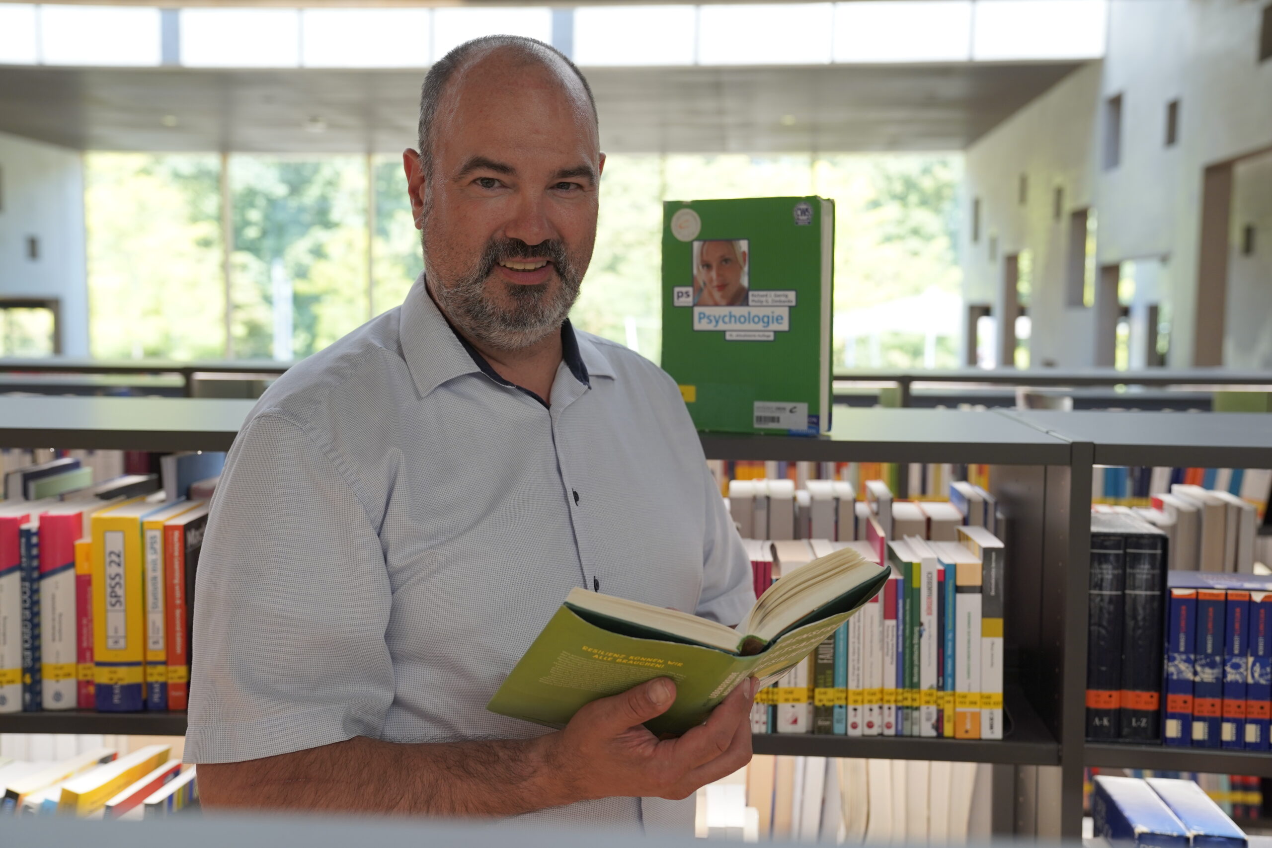 Ein bärtiger Mann steht in einer Bibliothek der Hochschule Coburg, hält ein aufgeschlagenes Buch in der Hand und lächelt. Umgeben von Bücherregalen voller bunter Bücher zu Themen wie Gesundheitswissenschaften und Digital Health genießt er das warme Sonnenlicht, das durch die großen Fenster hereinströmt. at Hochschule Coburg