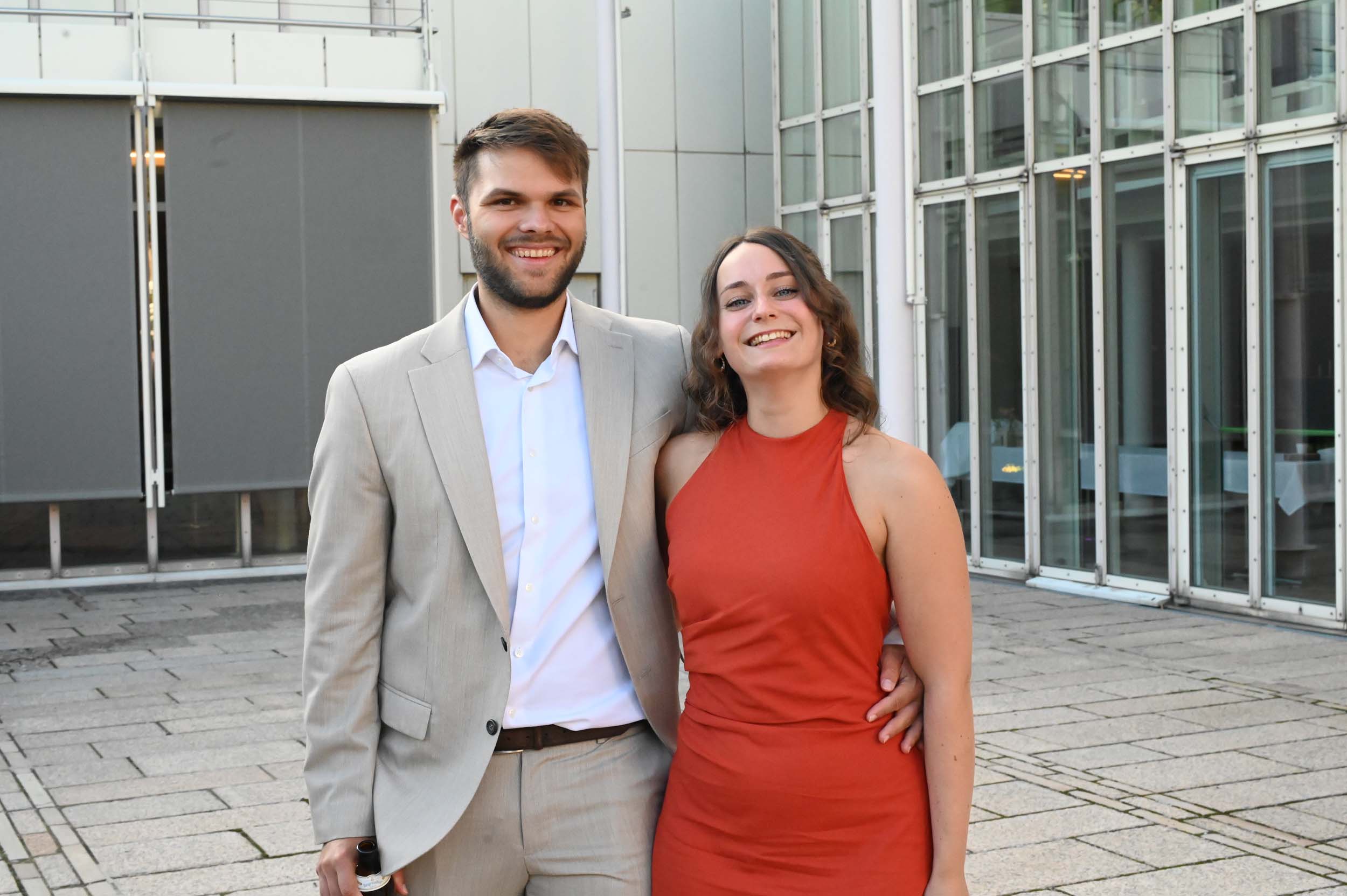 Ein lächelnder Mann in einem beigen Anzug steht neben einer Frau in einem roten, ärmellosen Kleid. Diese Absolventen posieren gemeinsam im Freien auf einem gepflasterten Platz vor einem modernen Glasgebäude und feiern ihren Abschied in fröhlicher Kameradschaft. at Hochschule Coburg