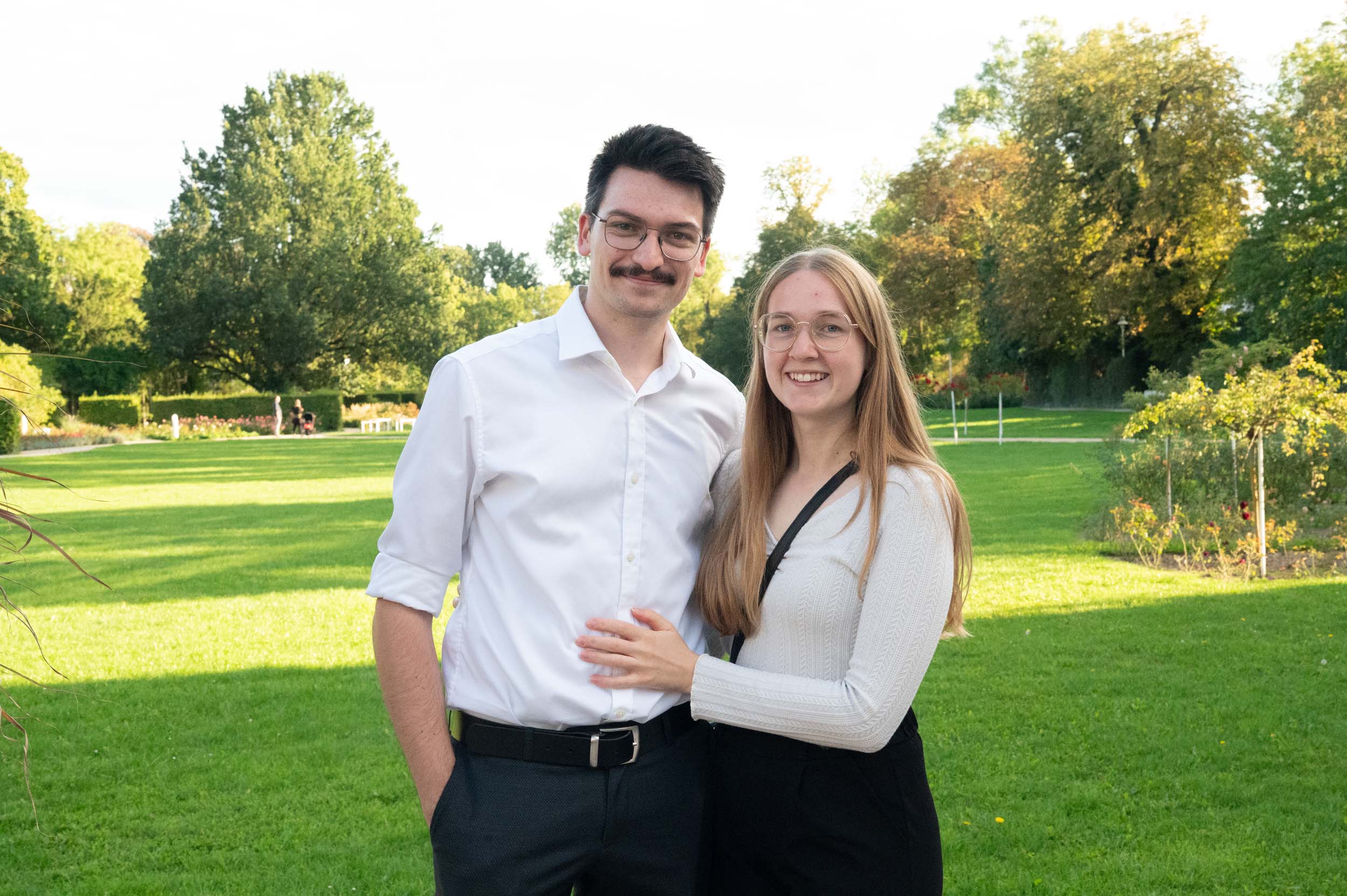 Ein lächelndes Paar steht gemeinsam auf einer Rasenfläche, im Hintergrund sind Bäume und Sonnenlicht zu sehen. Der Mann trägt ein weißes Hemd und eine Brille, die Frau trägt eine Brille und einen hellen Pullover. Das Bild fängt einen freudigen Abschiedsmoment ein, in dem die Absolventen ihre Erfolge feiern. at Hochschule Coburg