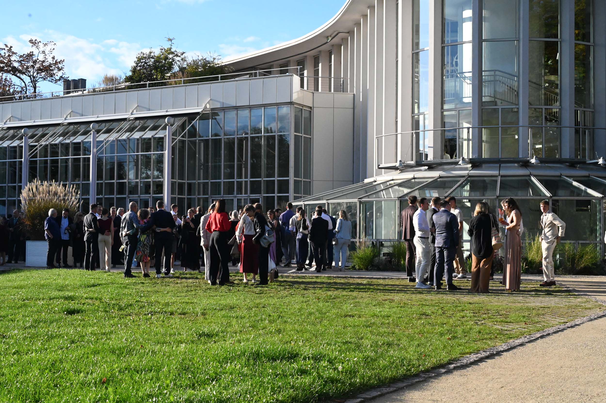 Eine Gruppe von Absolventinnen und Absolventen in halbformeller Kleidung hat sich an einem sonnigen Tag vor einem modernen Glasgebäude versammelt und unterhält sich auf einer Rasenfläche und einer Terrasse, während sie ihren Abschied feiern. at Hochschule Coburg