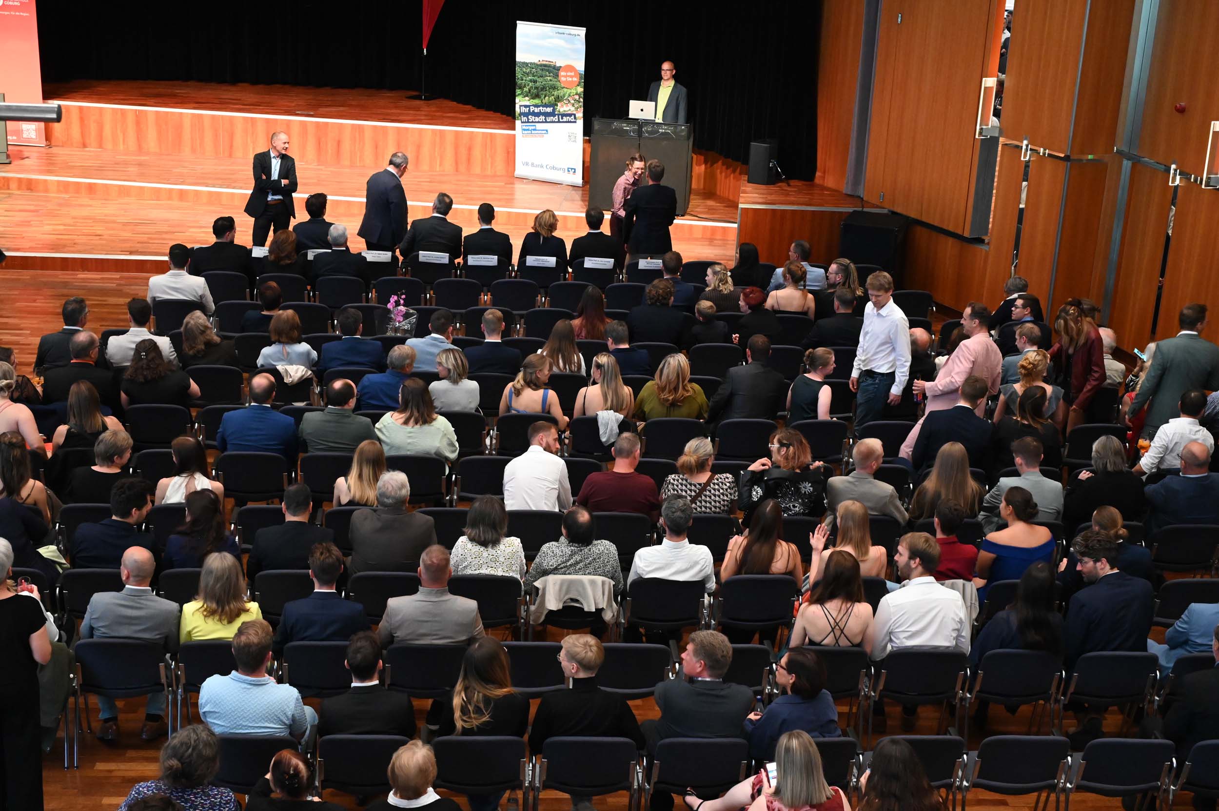 Ein großes Publikum, darunter viele Absolventen und Absolventinnen, sitzt in Stuhlreihen in einem Hörsaal und blickt auf eine Bühne, auf der eine Person am Rednerpult steht. Einige stehen und unterhalten sich, während andere sitzen und darauf warten, dass die Abschiedsfeier beginnt. at Hochschule Coburg