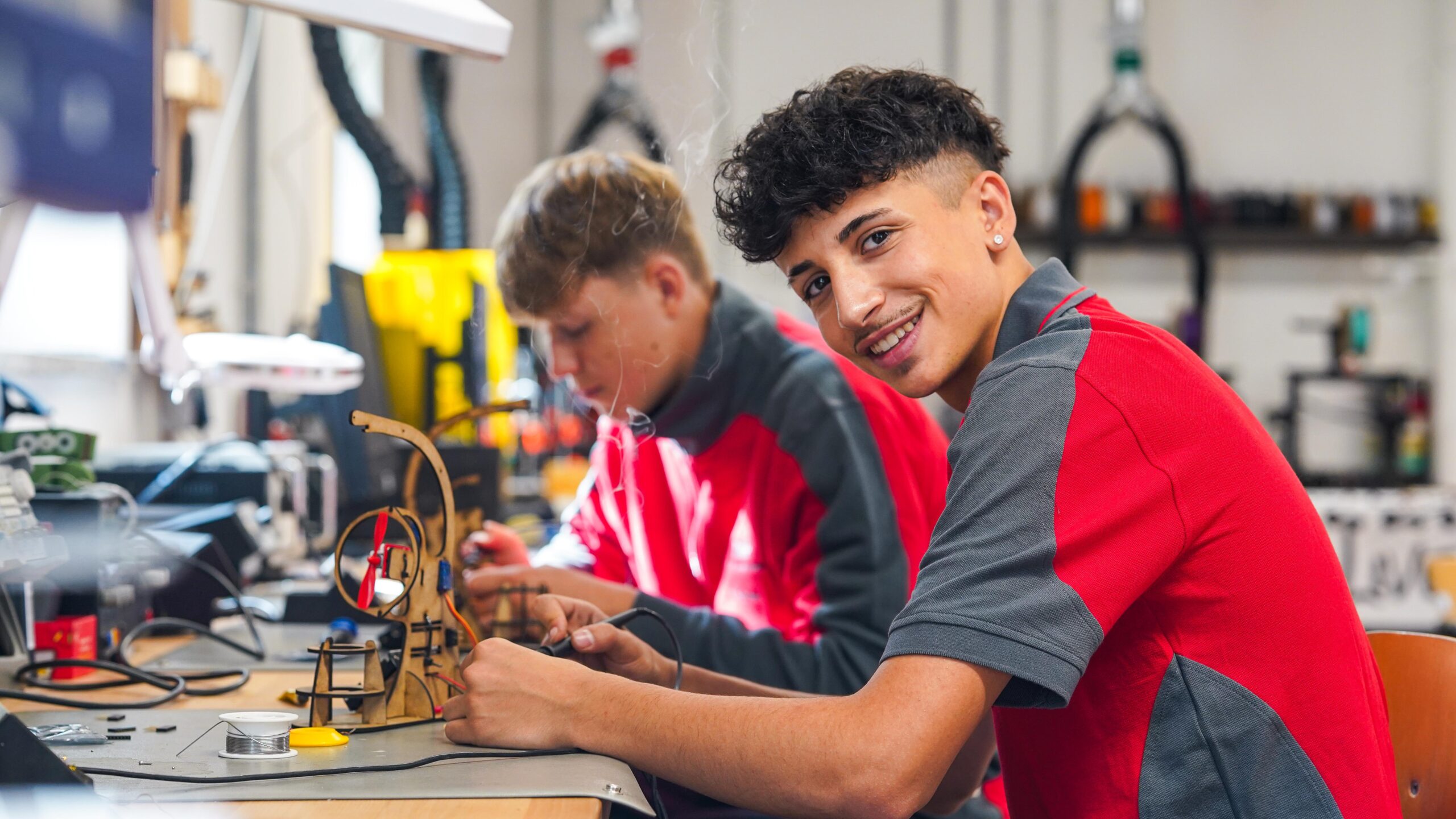 Zwei junge Menschen in roten und grauen Uniformen arbeiten an einer Werkbank an Elektronikprojekten und zeigen dabei echten Teamgeist. Der eine lächelt in die Kamera, während er ein Gerät zusammenbaut, der andere konzentriert sich im Hintergrund auf das Löten. at Hochschule Coburg