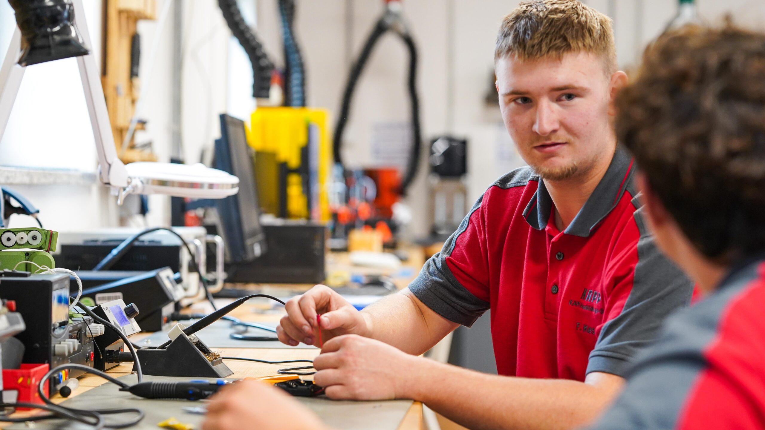 Zwei Personen in roten und grauen Arbeitsuniformen sitzen an einer Werkbank mit elektronischen Geräten und unterhalten sich in einer Werkstatt oder einem technischen Labor. Ihr Zusammenspiel spiegelt gelebte Zusammenarbeit wider und zeigt, wie der Teamgeist im CREAPOLIS Makerspace gestärkt wird. at Hochschule Coburg