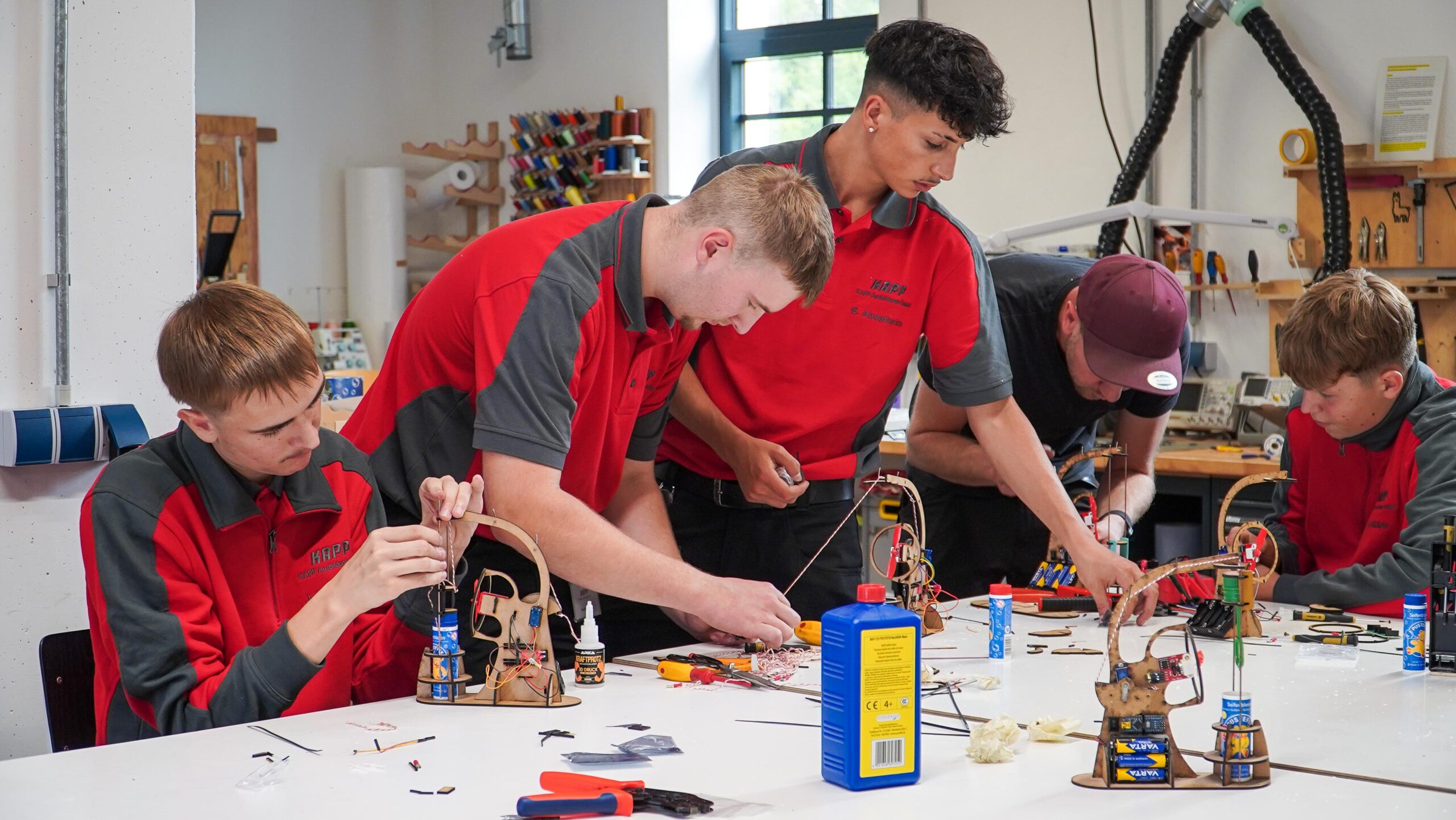 Fünf Jugendliche in roten und grauen Uniformen arbeiten an einem vollgestopften Tisch im CREAPOLIS Makerspace an kleinen Technik- oder Robotikprojekten und verwenden dabei Werkzeuge und elektronische Bauteile. Ihre Zusammenarbeit trägt dazu bei, den Teamgeist zu stärken, da sie in dieser kreativen Werkstattumgebung eng zusammenarbeiten. at Hochschule Coburg