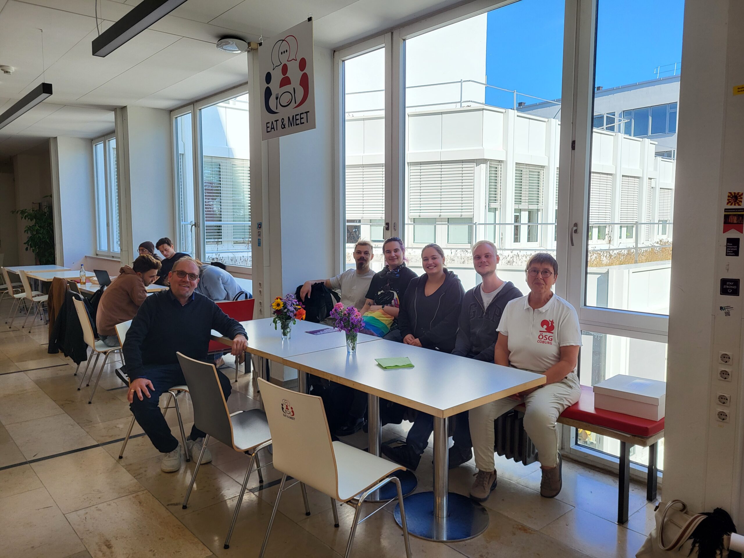 Bei einer „Eat & Meet“-Veranstaltung sitzt eine Gruppe von sechs Personen in einem hellen, modernen Raum um einen Tisch herum. at Hochschule Coburg