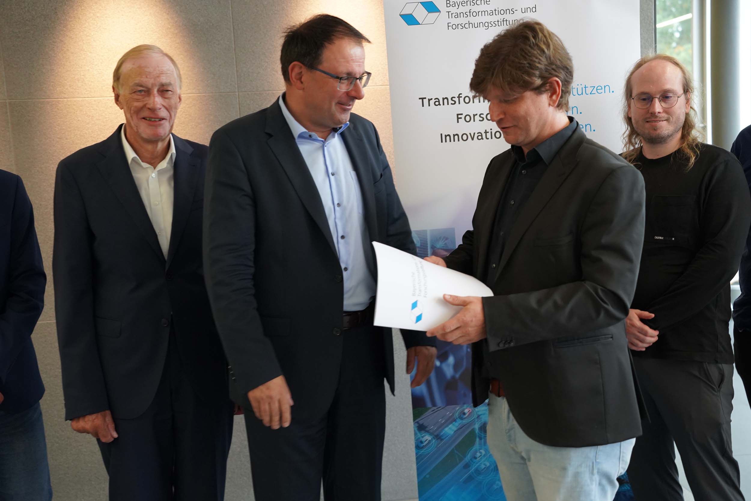 Ein Mann in einem schwarzen Blazer überreicht einem anderen Mann in einem dunklen Anzug vor einem Banner von „Coburger Forschung“ ein Papierdokument, während zwei weitere Männer neben ihnen stehen. at Hochschule Coburg