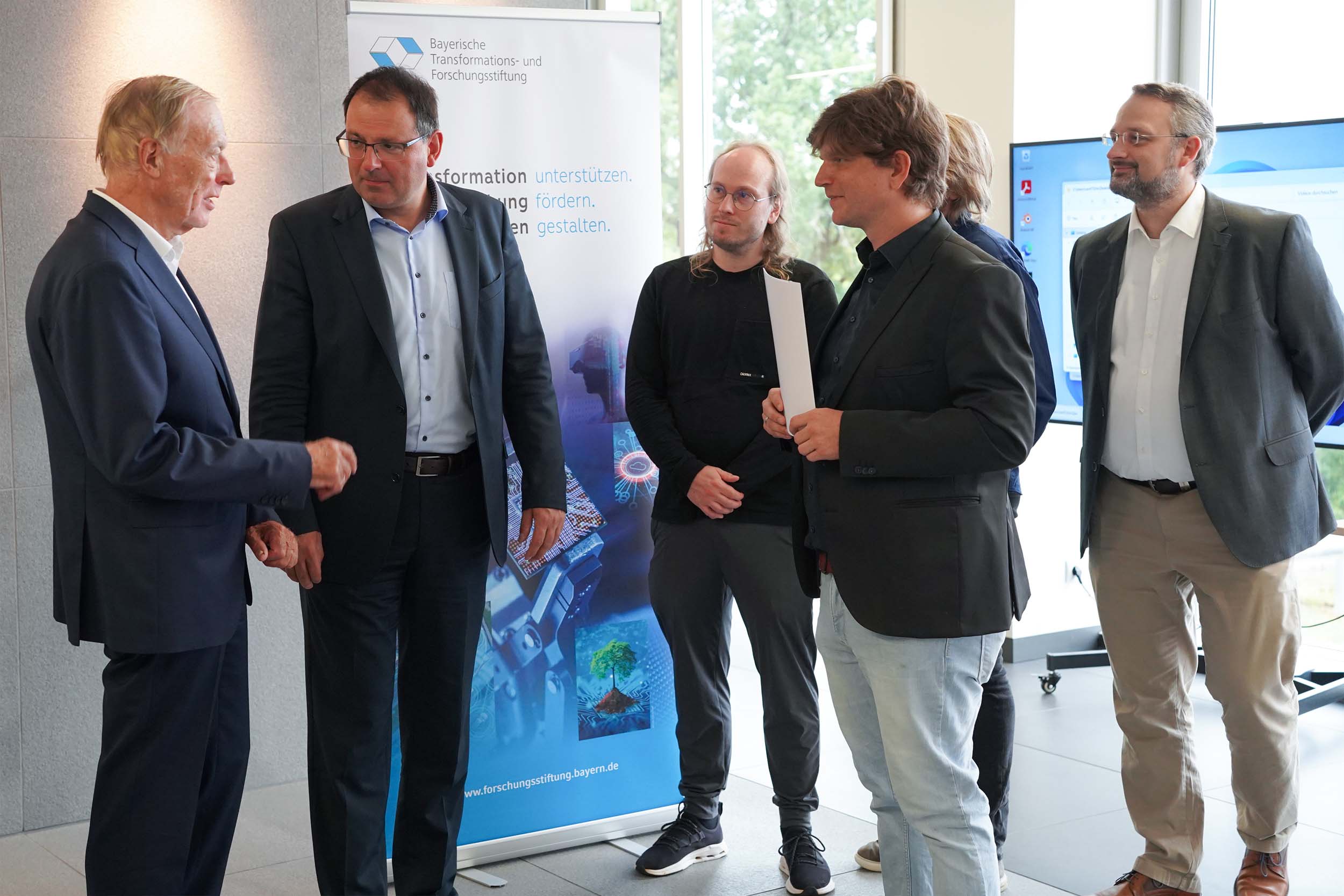 Fünf Männer in Business-Casual-Kleidung besprechen zukunftsorientierte Projekte der Coburger Forschung in der Nähe eines Werbebanners der Bayerischen Transformations- und Forschungsstiftung. at Hochschule Coburg