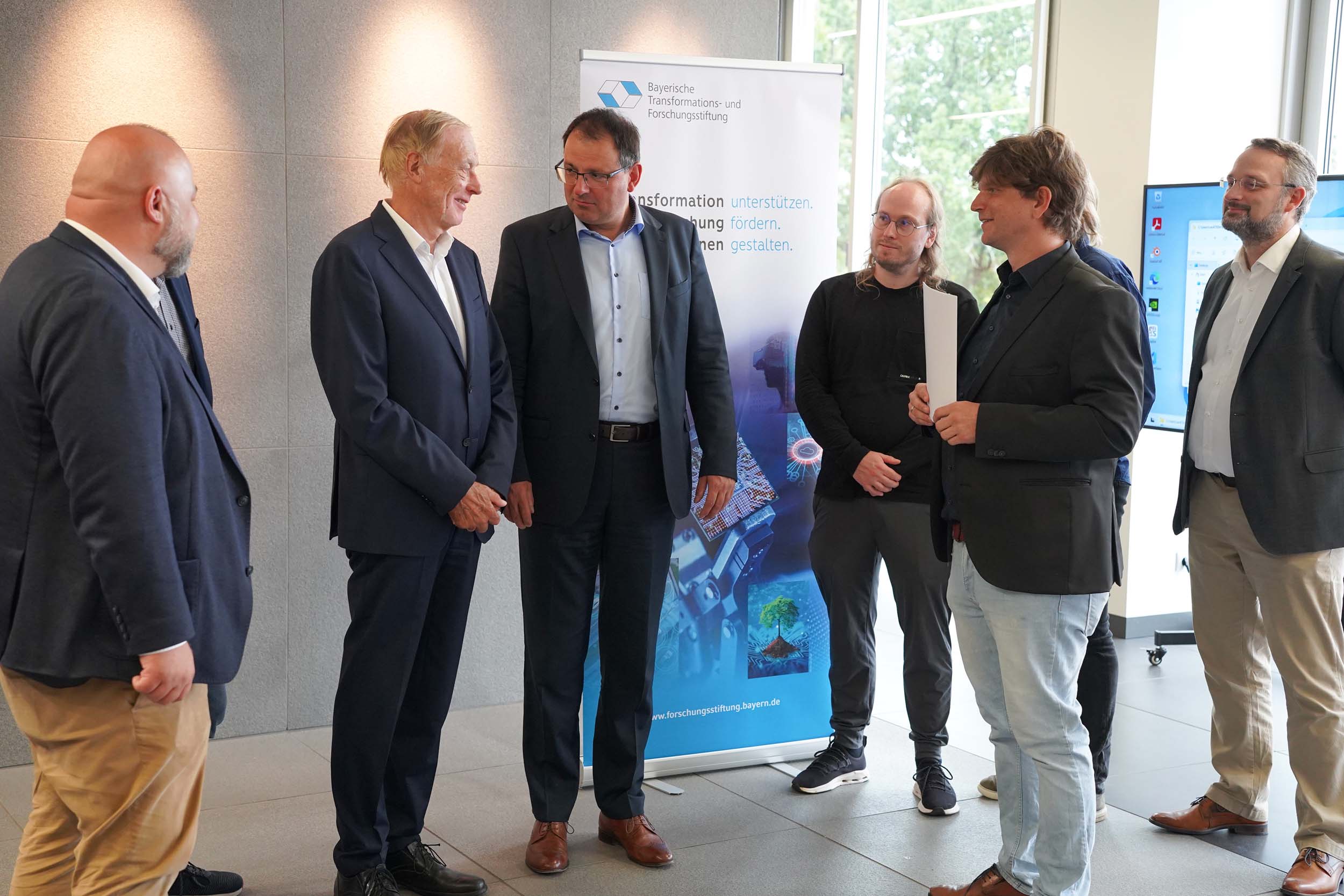 Sechs Männer stehen in einem Büroraum; einer von ihnen hält ein Dokument in der Hand, im Hintergrund ist ein Banner der „Coburger Forschung“ zur Zukunft der Computergrafik zu sehen. at Hochschule Coburg