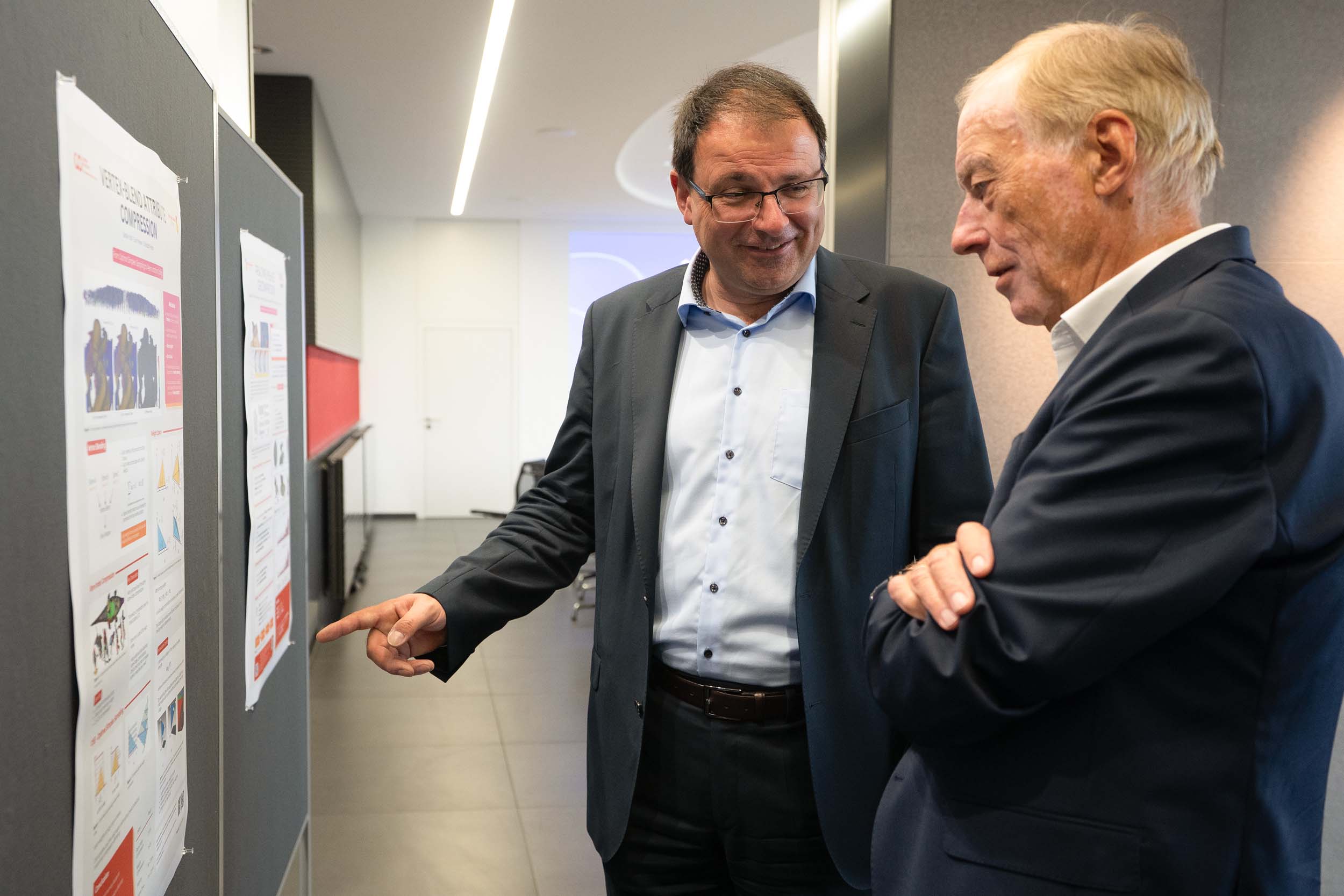 Zwei Männer in Geschäftskleidung betrachten ein an der Wand hängendes Forschungsplakat des Coburger Forschungsinstituts zum Thema Computergrafik. at Hochschule Coburg