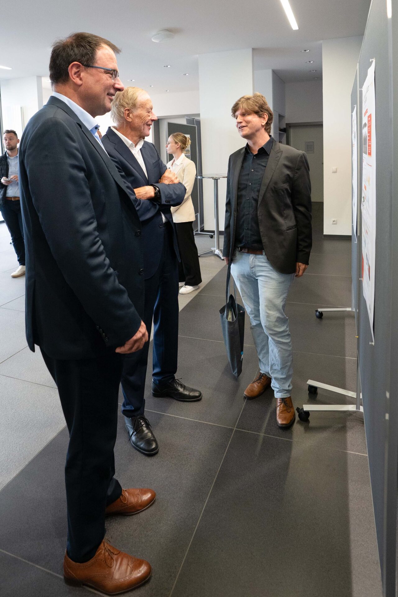Drei Männer in Geschäftskleidung stehen in der Lobby eines Büros und betrachten ein Plakat über die Coburger Forschung und Computergrafik. at Hochschule Coburg
