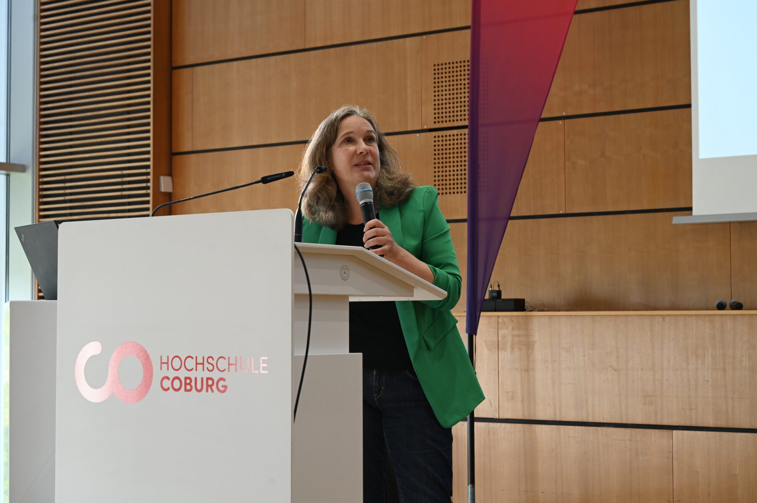 Eine Frau in einem grünen Blazer spricht an einem Rednerpult der Hochschule Coburg in ein Mikrofon. at Hochschule Coburg