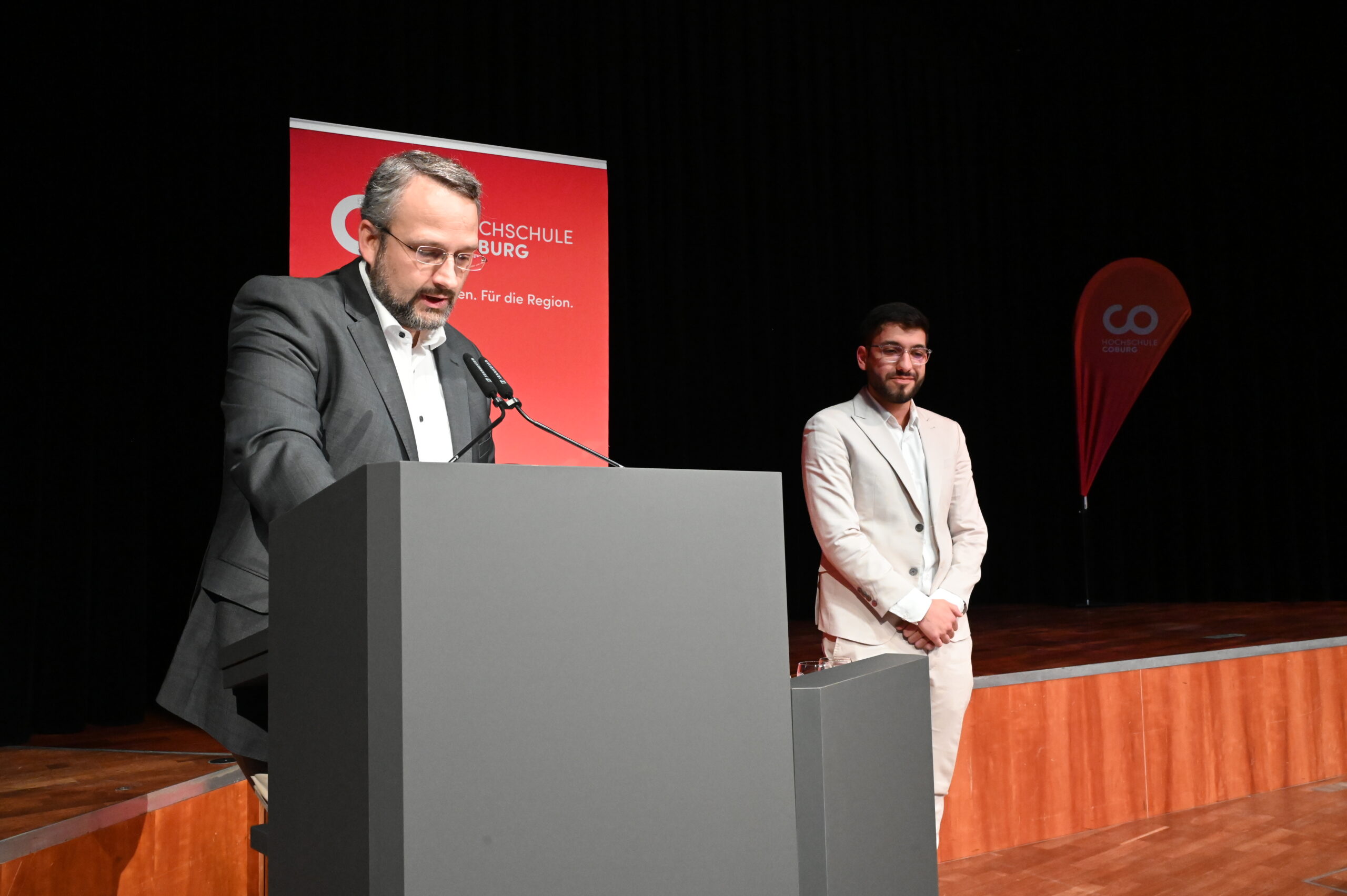 Ali Ali Mahmoud hält eine Rede am Rednerpult, während er auf einer Bühne mit Bannern der Hochschule Coburg den DAAD-Preis entgegennimmt. at Hochschule Coburg