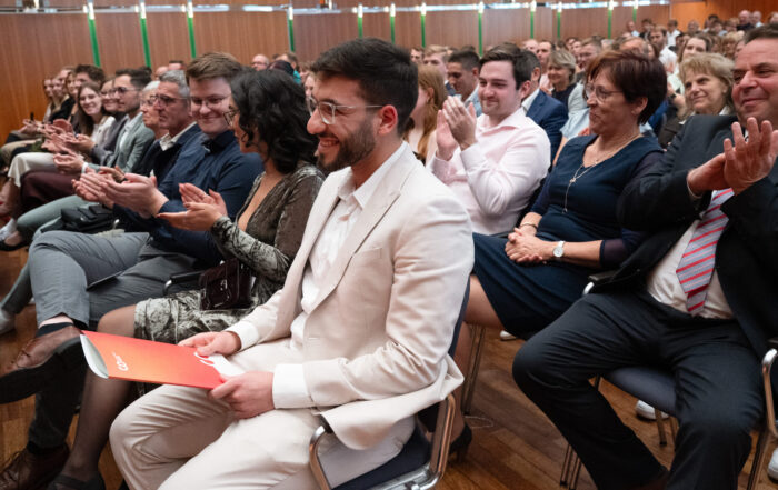 In einem mit Holz vertäfelten Raum applaudiert das Publikum, während Ali Ali Mahmoud, der einen hellen Anzug trägt und ein rotes DAAD-Preis-Programm in der Hand hält, im Vordergrund steht. at Hochschule Coburg