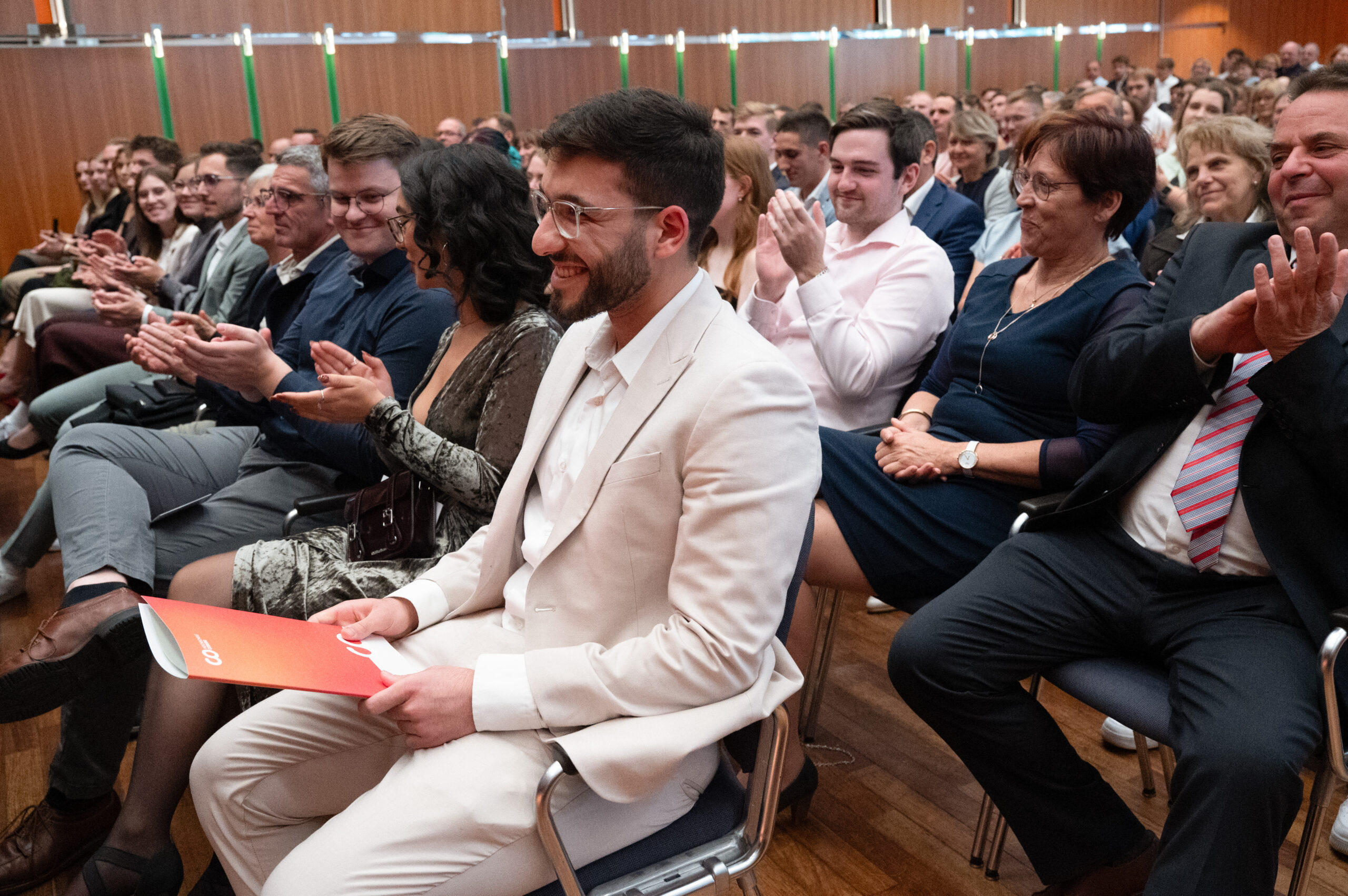 In einem mit Holz vertäfelten Raum applaudiert das Publikum, während Ali Ali Mahmoud, der einen hellen Anzug trägt und ein rotes DAAD-Preis-Programm in der Hand hält, im Vordergrund steht. at Hochschule Coburg