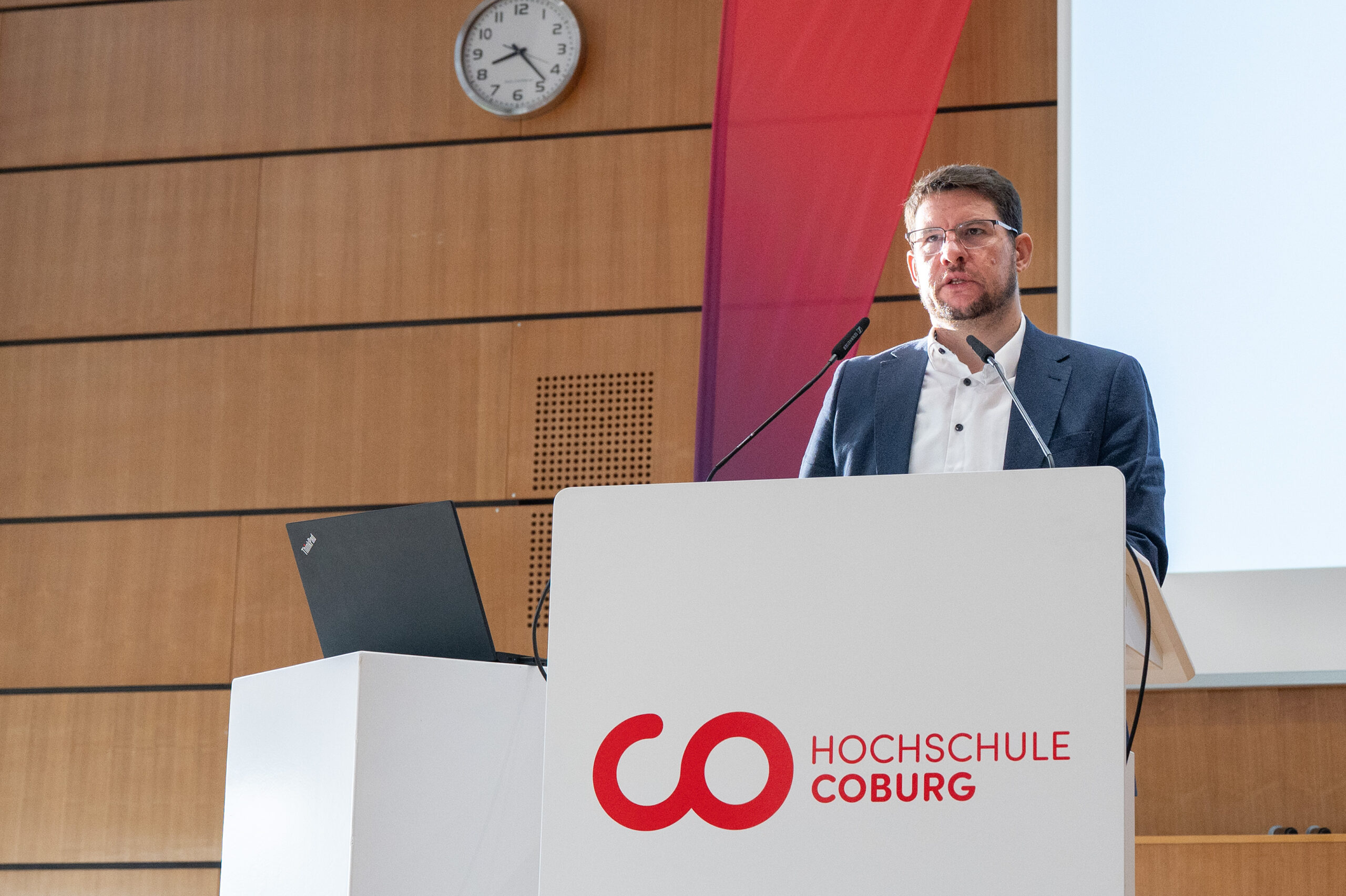 Ein Mann im Anzug und mit Brille hält während einer Präsentation zum Thema „Studieren in Coburg“ eine Rede an einem Rednerpult, das mit dem Logo der Hochschule Coburg versehen ist. at Hochschule Coburg