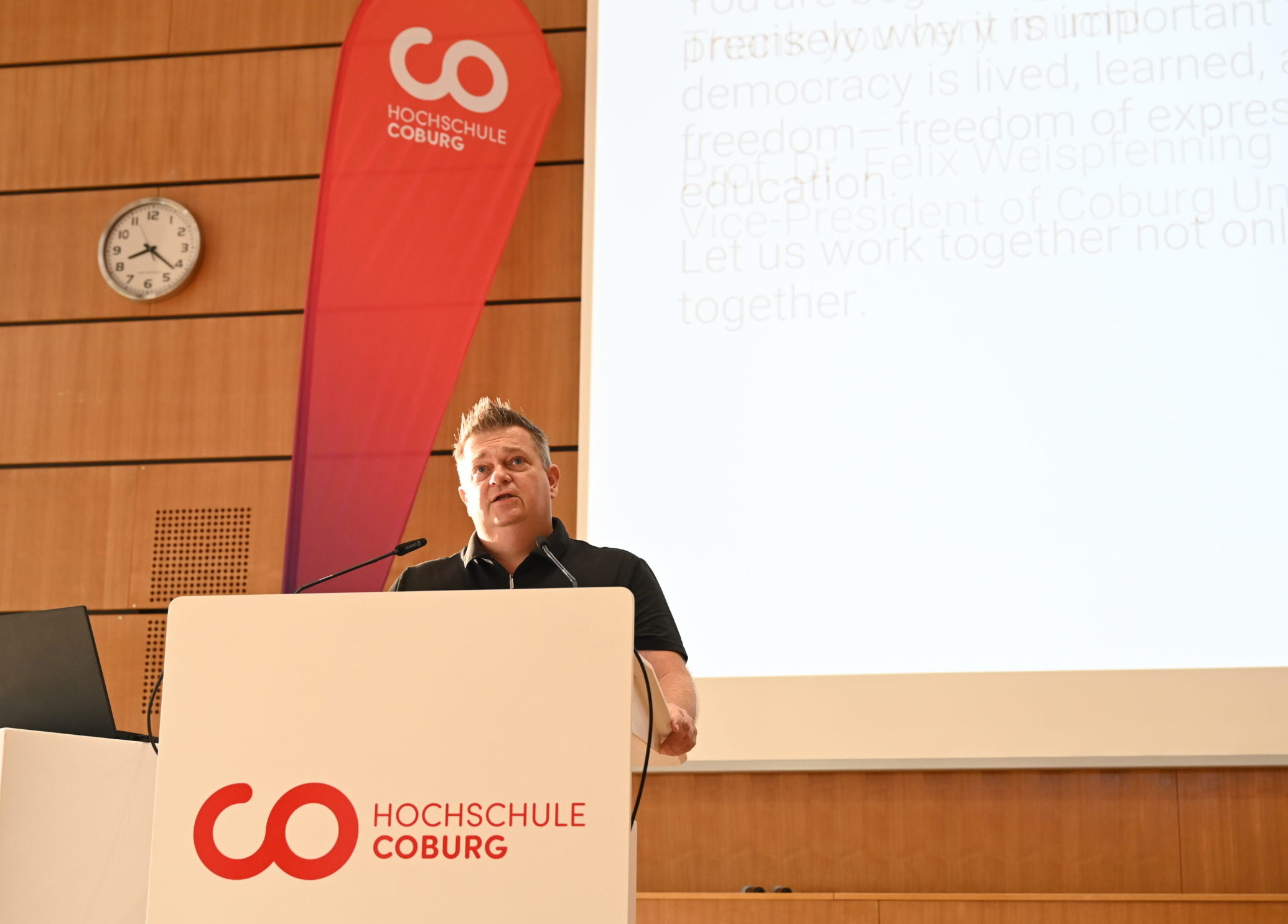 Ein Mann spricht während einer Veranstaltung zum Thema „Studieren in Coburg“ vor einer Leinwand von einem Rednerpult aus, das mit dem Logo der Hochschule Coburg versehen ist. at Hochschule Coburg