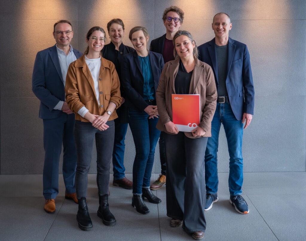 Sechs Kollegen aus dem Bereich der klinischen Sozialarbeit lächeln gemeinsam in einer Büro-Lobby, wobei eine Person während der Jubiläumsfeier eine orangefarbene Mappe in der Hand hält. at Hochschule Coburg