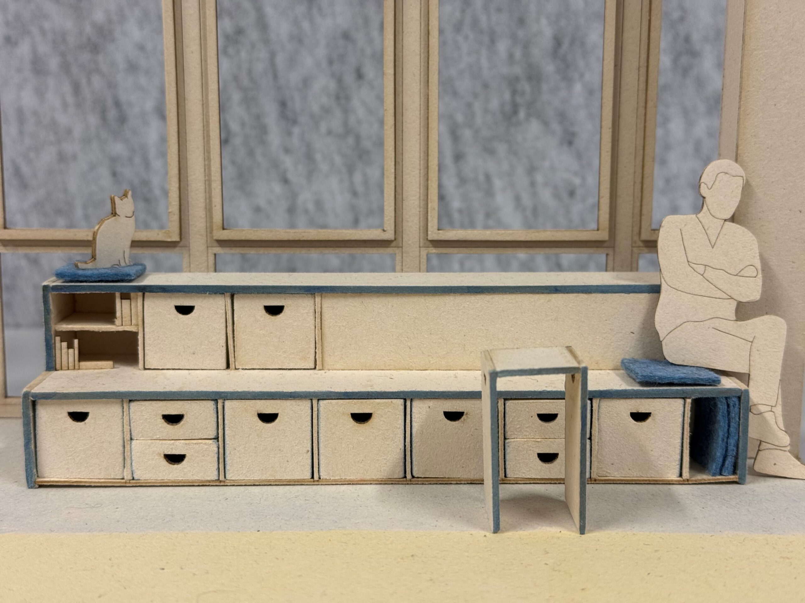 Ein maßstabsgetreues Modell eines Einbaumöbels mit mehreren Schubladen, einem kleinen Hocker, einer Katzenfigur und einem ausgeschnittenen Bild einer Person, die am Ende sitzt. at Hochschule Coburg