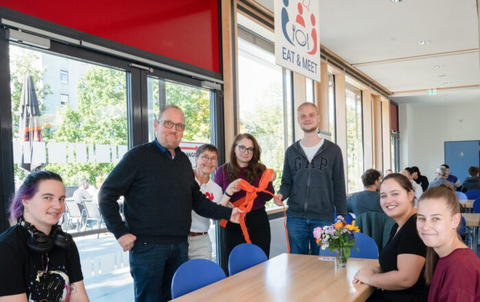 Sechs Personen stehen unter einem „Eat & Meet“-Schild um einen Tisch herum und halten ein orangefarbenes Band in den Händen, das in einem hellen Speiseraum einen Raum für Design-Automatisierung symbolisiert. at Hochschule Coburg