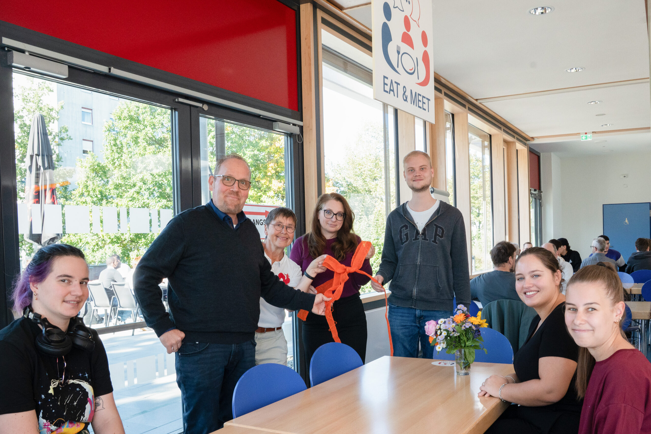 Sechs Personen stehen unter einem „Eat & Meet“-Schild um einen Tisch herum und halten ein orangefarbenes Band in den Händen, das in einem hellen Speiseraum einen Raum für Design-Automatisierung symbolisiert. at Hochschule Coburg