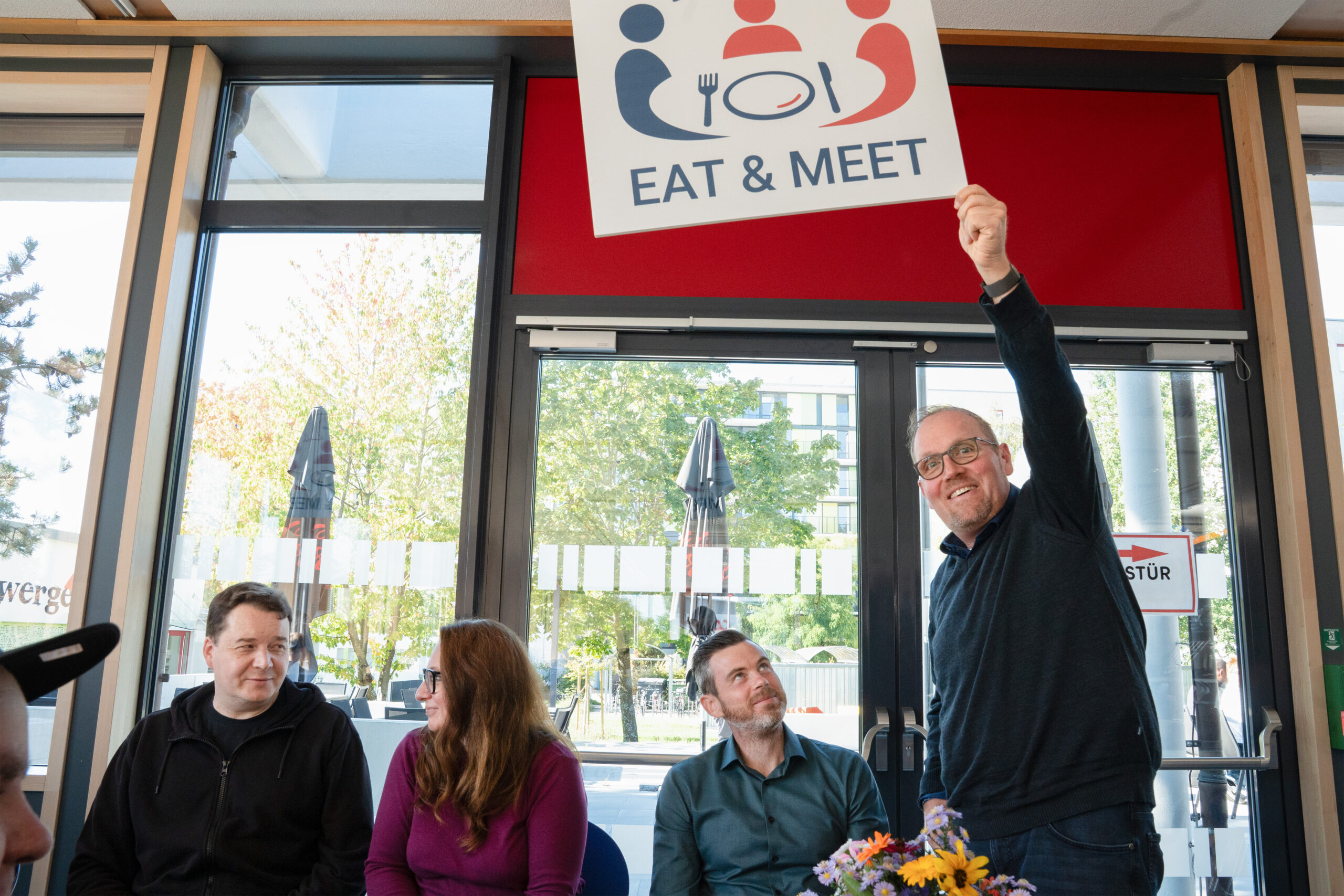 Ein Mann hält ein Schild mit der Aufschrift „EAT & MEET“ über drei Personen an einem Tisch und unterstreicht damit das automatisierte Konzept der Veranstaltung. at Hochschule Coburg