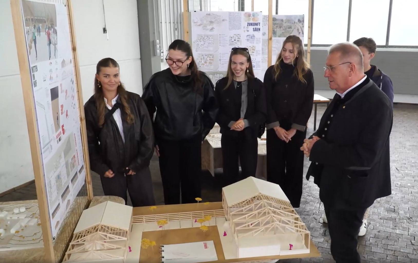 Ein Mann in einer schwarzen Jacke bespricht ein architektonisches Semesterprojekt mit vier jungen Frauen, die vor einem maßstabsgetreuen Modell und Projektplakaten stehen. at Hochschule Coburg