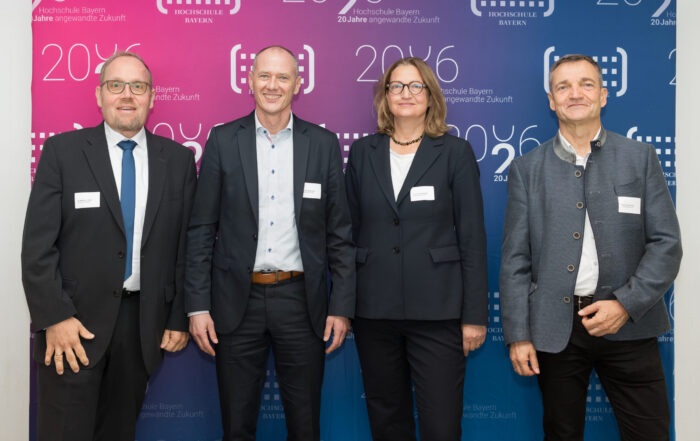 Vier Personen stehen in Coburg in einer Reihe vor einer blau-lila Jubiläums-Kulisse der Hochschule Bayern. at Hochschule Coburg