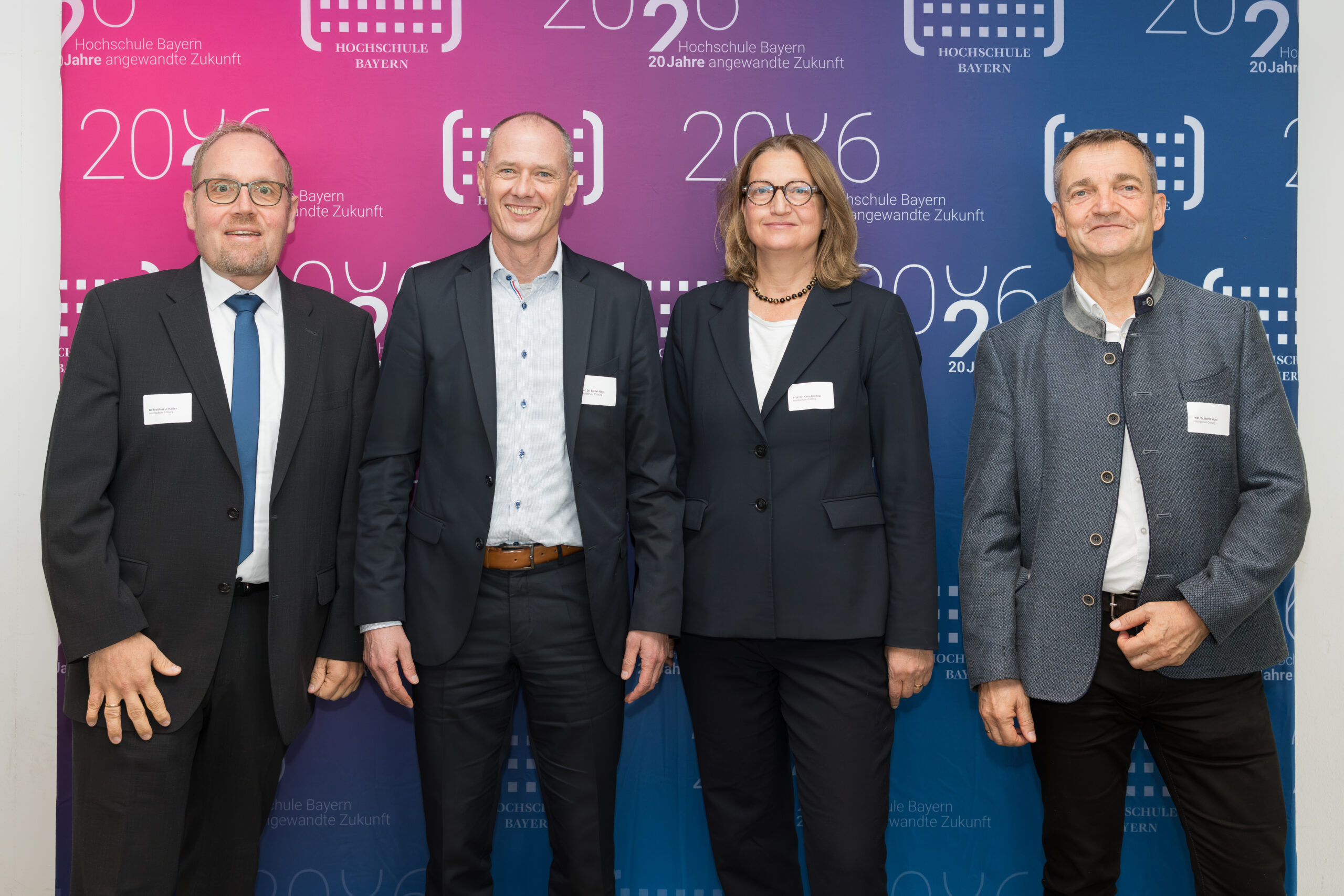 Vier Personen stehen in Coburg in einer Reihe vor einer blau-lila Jubiläums-Kulisse der Hochschule Bayern. at Hochschule Coburg