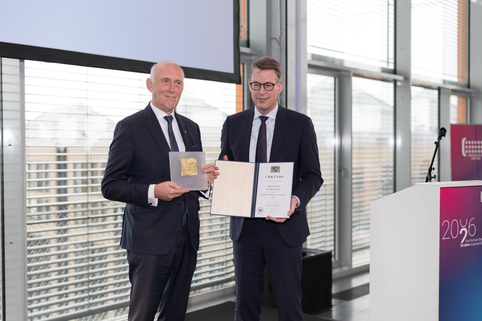Zwei Männer in Anzügen stehen während einer Jubiläumsfeier der Hochschule Bayern auf einem Podium; der eine hält eine goldfarbene Plakette in der Hand, der andere eine Urkunde. at Hochschule Coburg