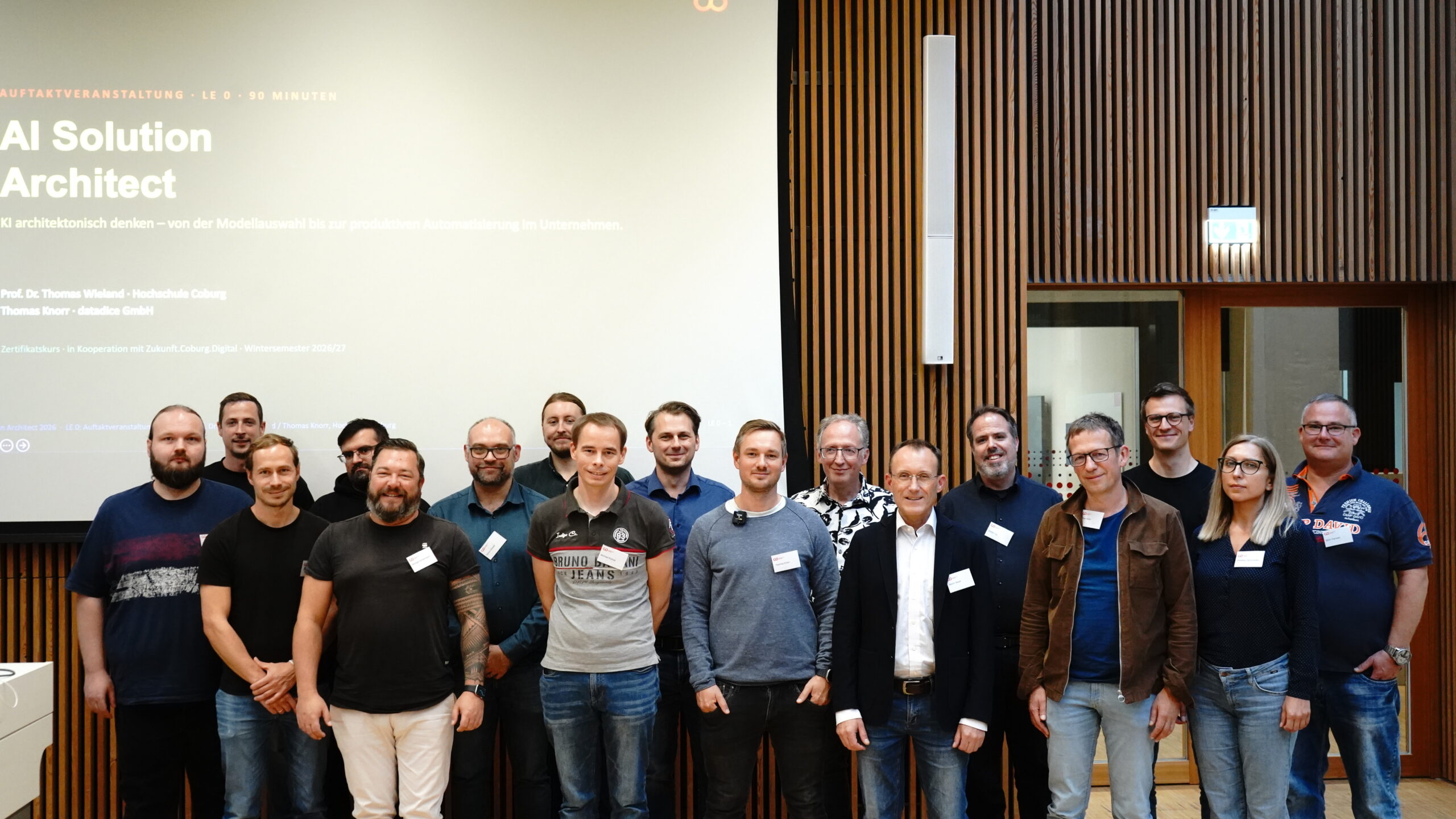 Eine Gruppe von siebzehn Personen, die in einer Reihe vor einer Präsentationswand mit der Aufschrift „AI Solution Architect“ stehen, die sich mit KI-Lösungen befasst. at Hochschule Coburg