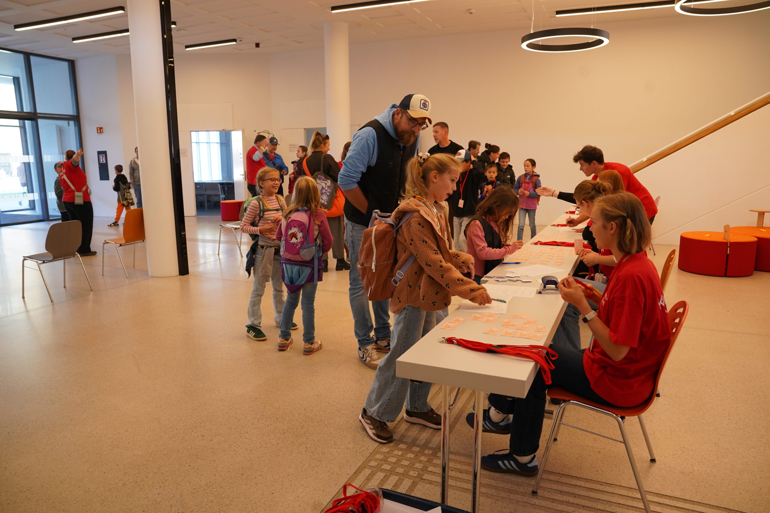 Kinder und Erwachsene versammeln sich um einen Anmeldetisch der KinderUni, an dem Menschen in roten T-Shirts rote Umhängebänder und Namensschilder an neue Teilnehmer und Stammgäste verteilen. at Hochschule Coburg