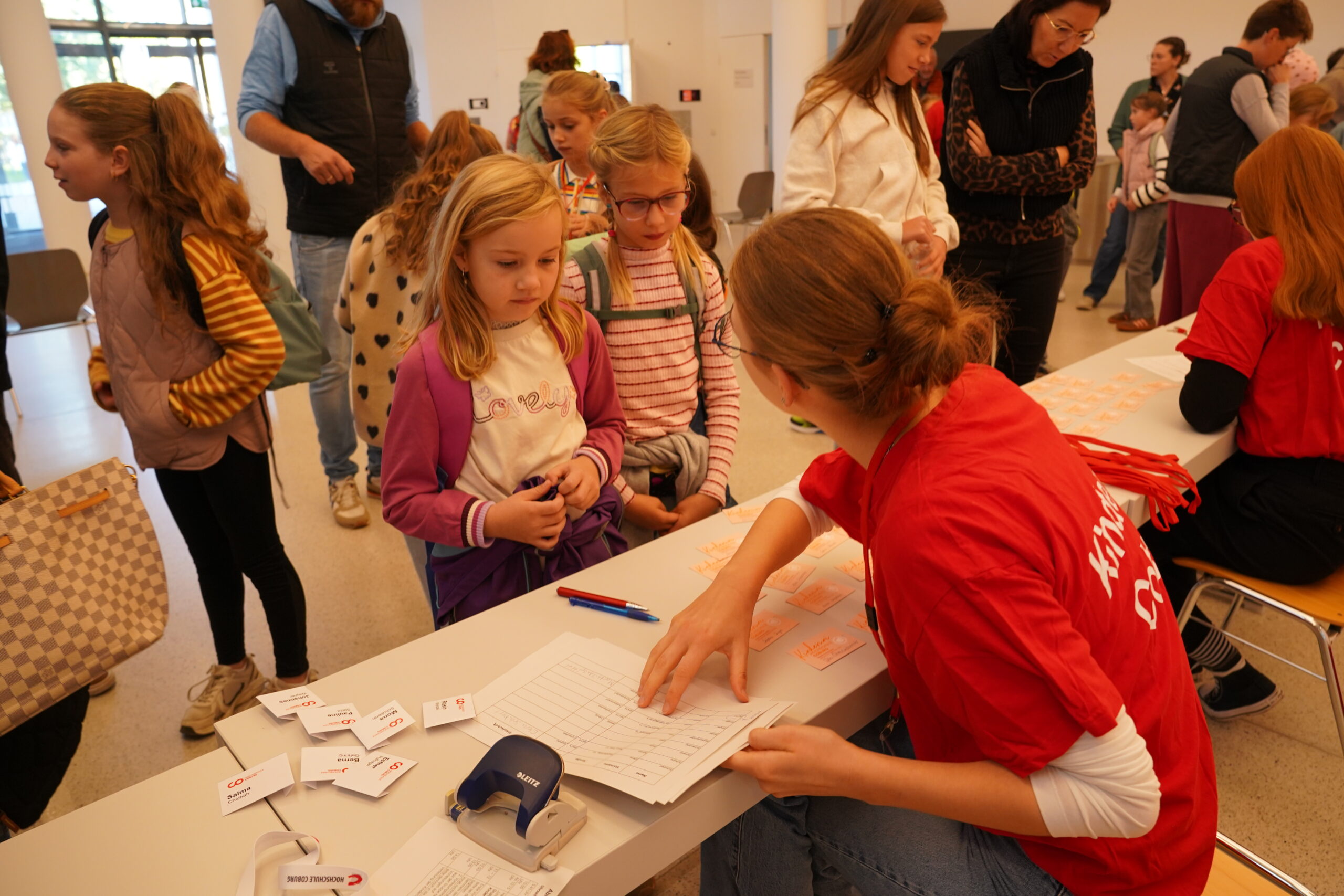 Ein ehrenamtlicher Helfer in einem roten T-Shirt registriert junge „KinderUni“-Teilnehmer an einem Anmeldeschalter und händigt ihnen Namensschilder und Unterlagen aus. at Hochschule Coburg
