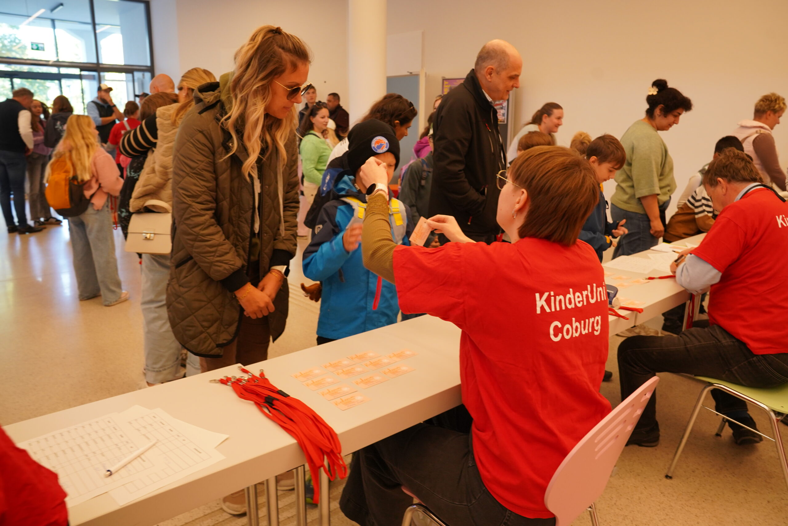 Ein Mitarbeiter in einem roten „KinderUni Coburg“-T-Shirt händigt einem Kind am Anmeldeschalter ein Namensschild aus und heißt die jungen Stammgäste wieder willkommen. at Hochschule Coburg